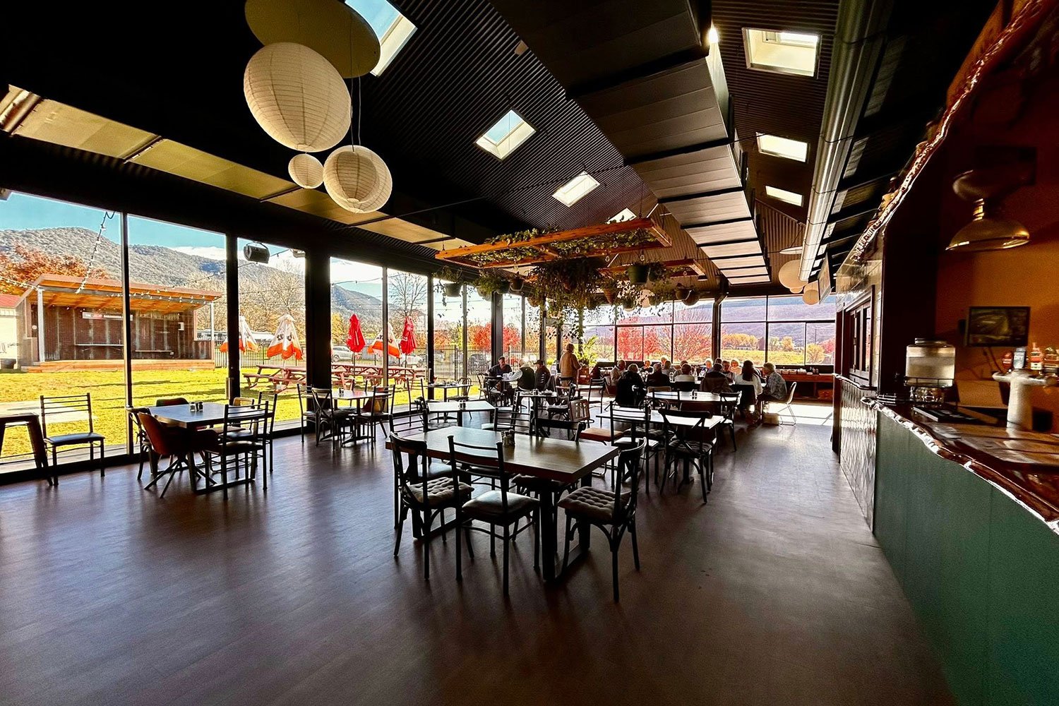 Interior of The Dederang Hotels Pavilion, with large windows showing mountains and fall foliage outside. The space has tables and chairs, lanterns and greenery decorations hanging from the ceiling.
