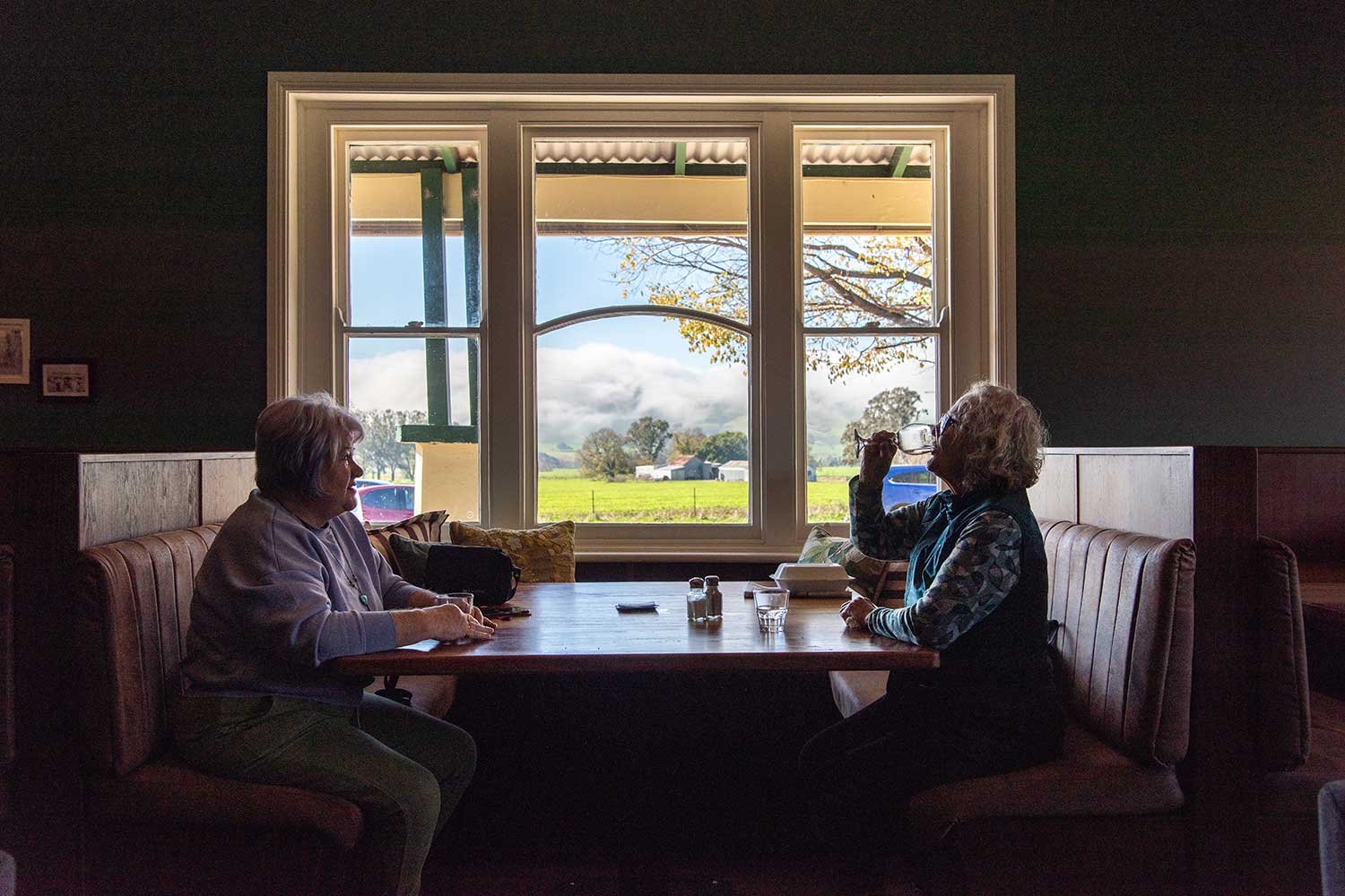 Two women sitting in one of The Dederang Hotel's cozy restaurant booths, facing each other, with a large window showing a scenic countryside view of fields, trees and a cloudy sky.