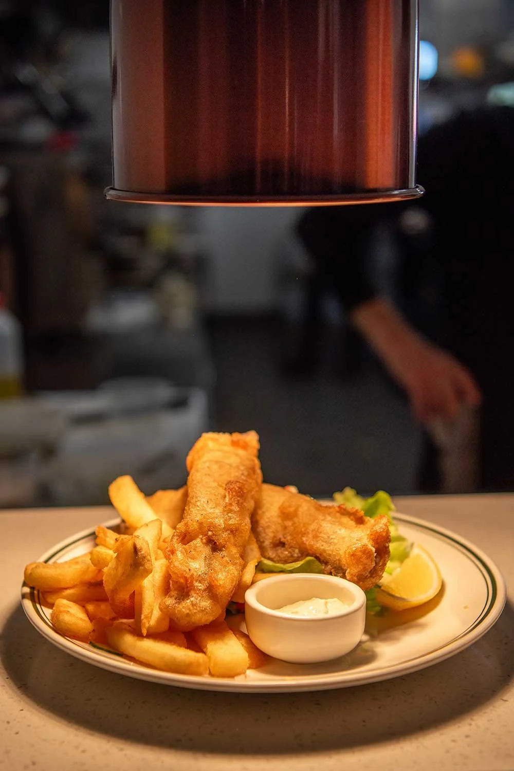 Plate of fish and chips with tartar sauce and lemon wedge, garnished with lettuce, under a heat lamp in The Dederang Hotel's kitchen.