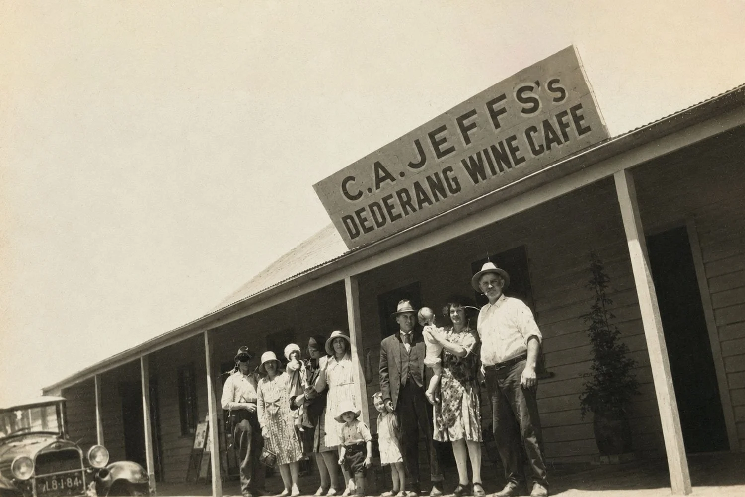 A group photograph taken circa 1950 showing men, women and children in vintage clothing, with a vintage car nearby in front of building with a sign that reads 'C. A. Jeffs Dederang Wine Cafe'.