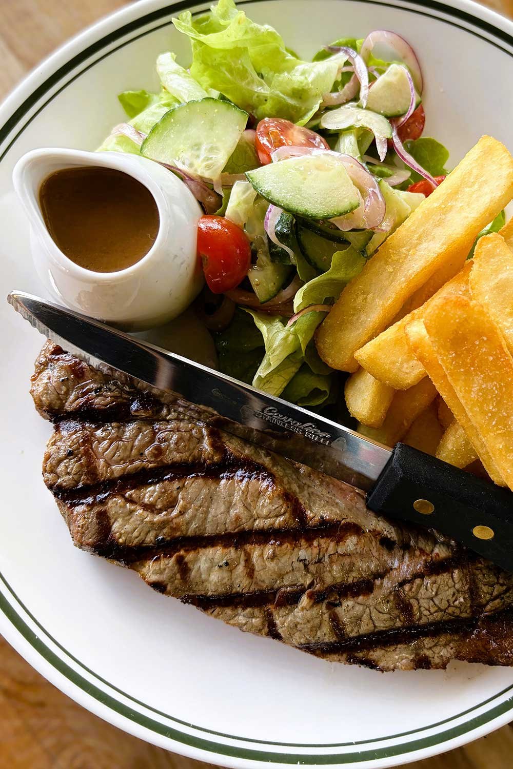 A plate of grilled steak with grill marks, a side salad with cucumbers, cherry tomatoes and lettuce, thick-cut chips and a small pitcher of brown gravy.
