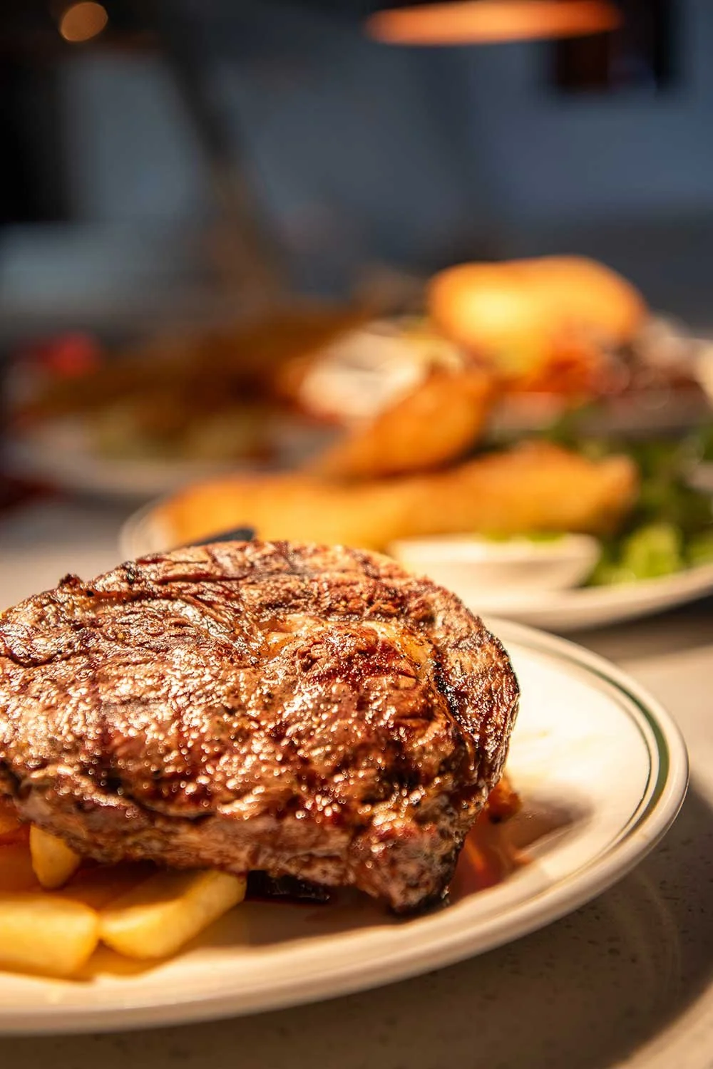 Close-up of a steak on a plate with fries underneath, with more food in the background.