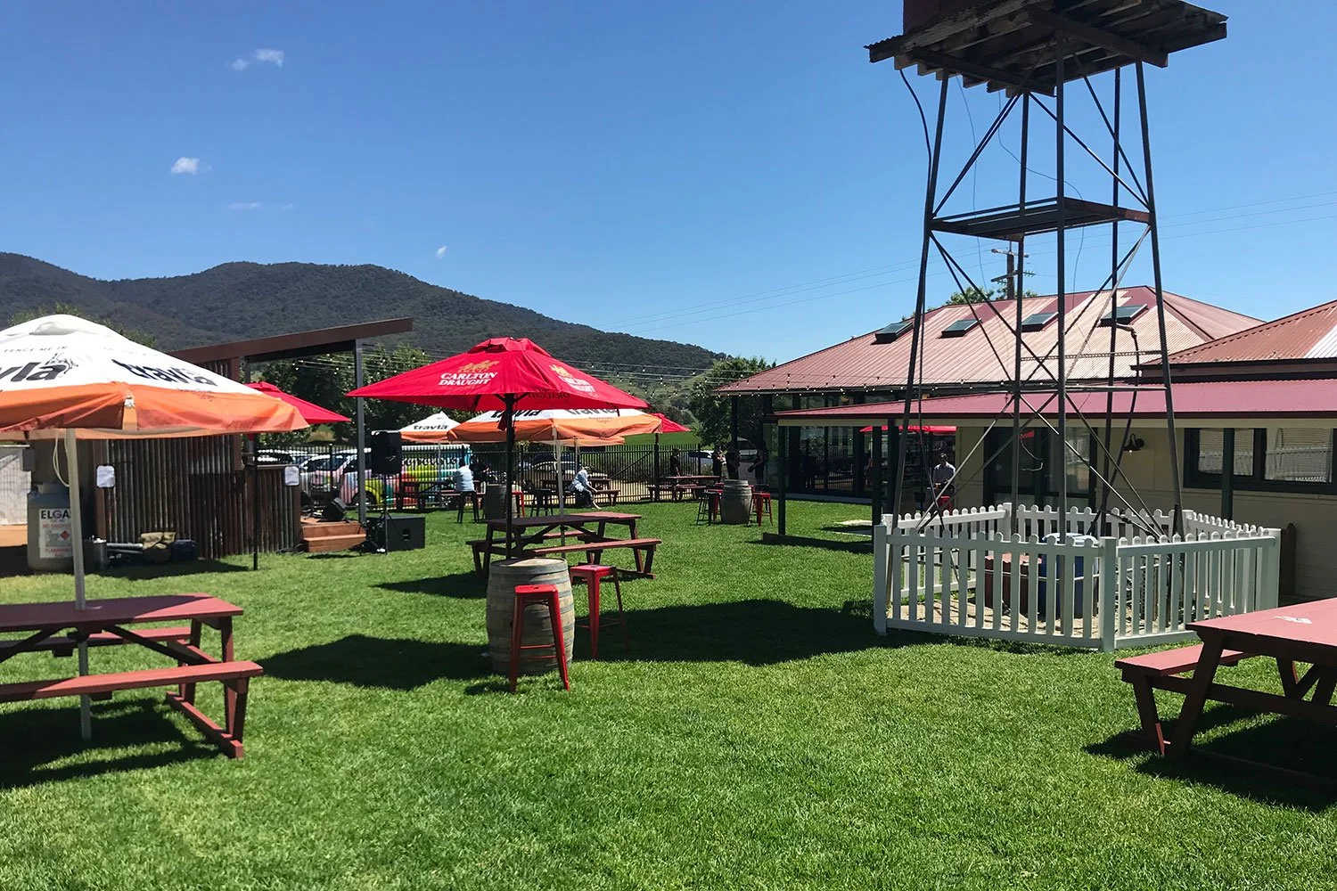 Outdoor seating area with red and orange umbrellas, picnic tables, a small fenced-in structure with a tower, green grass, building with a red roof, mountains in the background, and people in the distance.