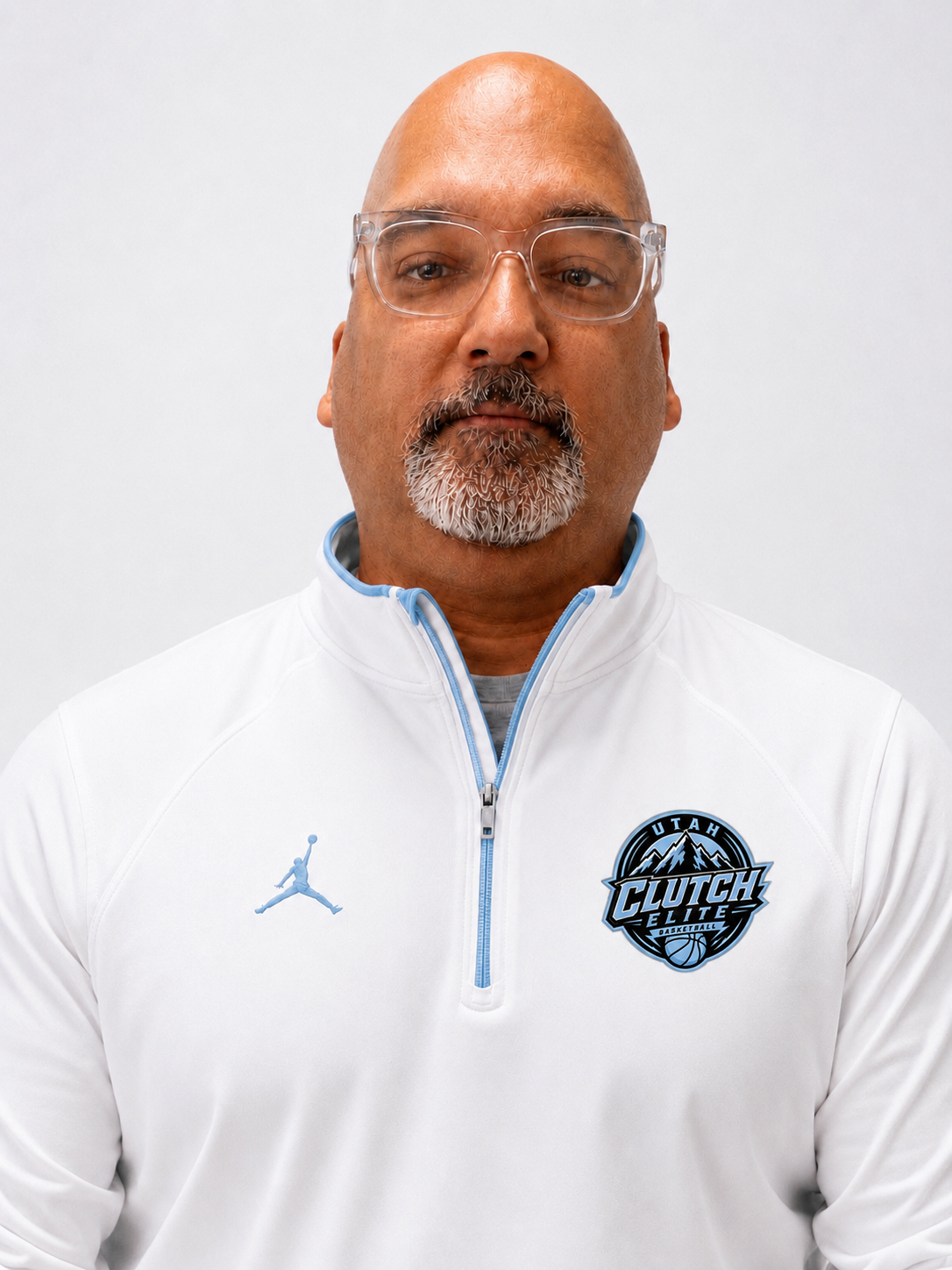 Portrait of a middle-aged man with glasses and a gray beard wearing a white sports jacket with Clutch Elite basketball logo and Jordan brand logo, against a plain white background.
