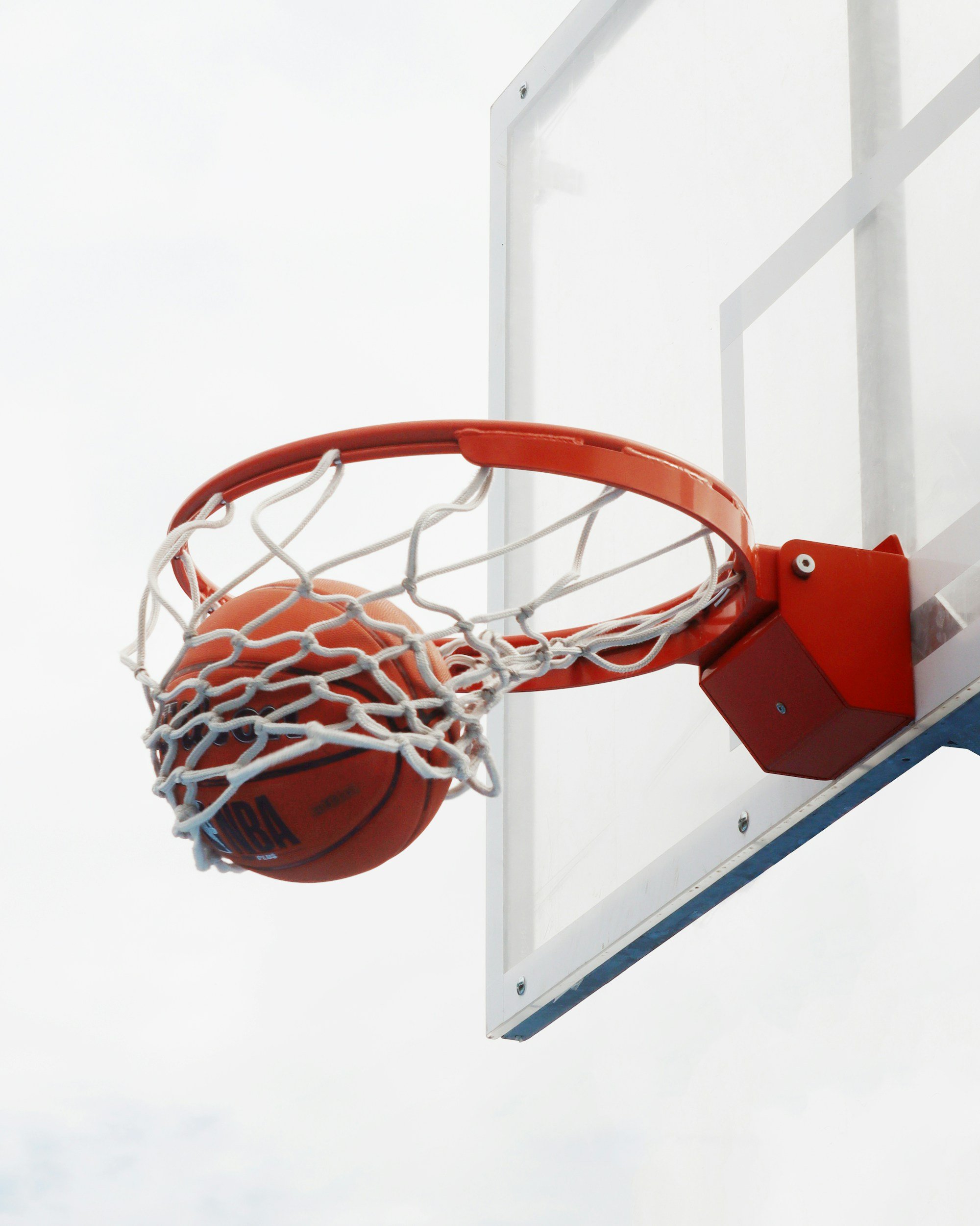 An orange basketball going through a basketball hoop mounted on a white backboard against a white sky.