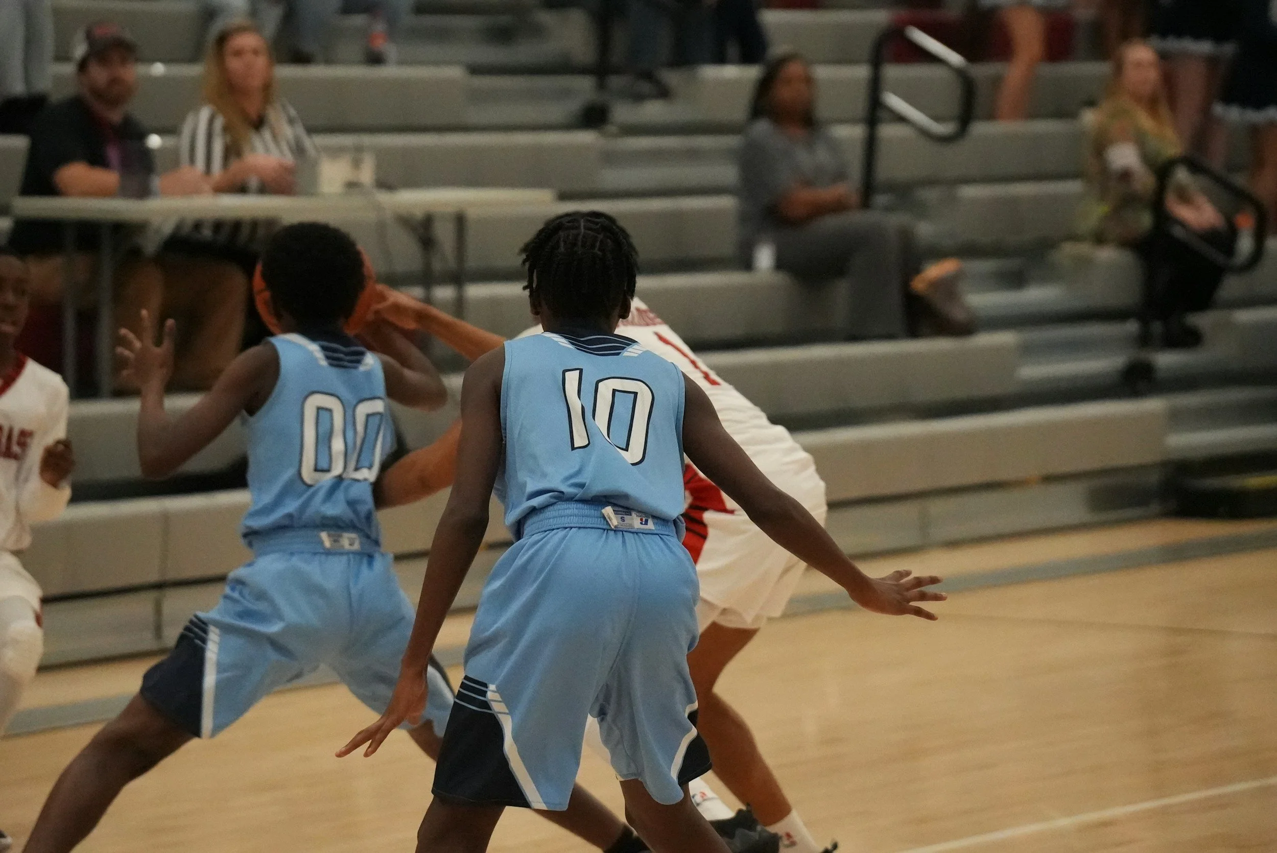 Young basketball players in blue uniforms actively playing on the court during a game, with spectators sitting in bleachers in the background.