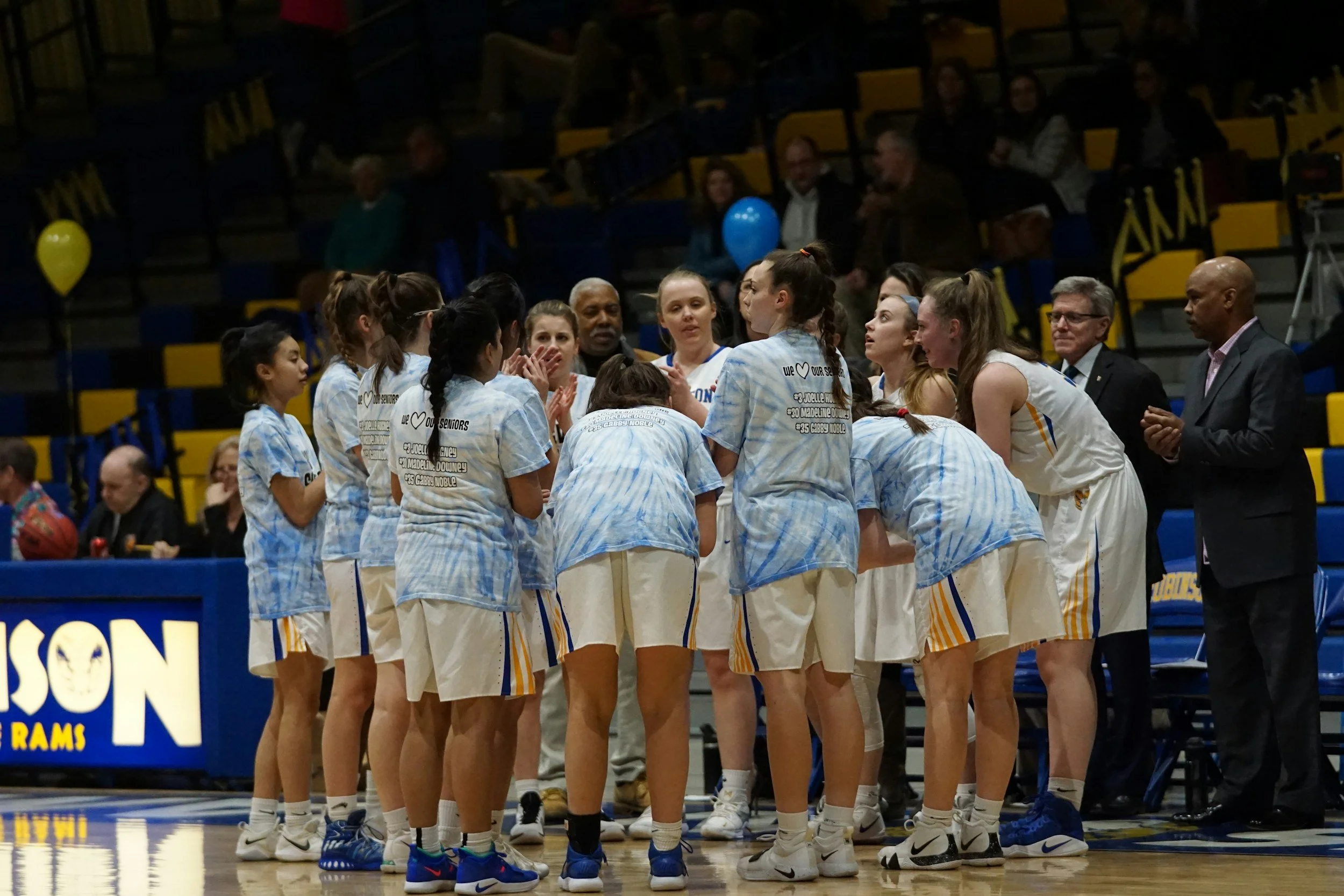 A group of female basketball players in white, yellow, and blue uniforms huddle together during a game or practice, appearing to listen to coaching instructions.