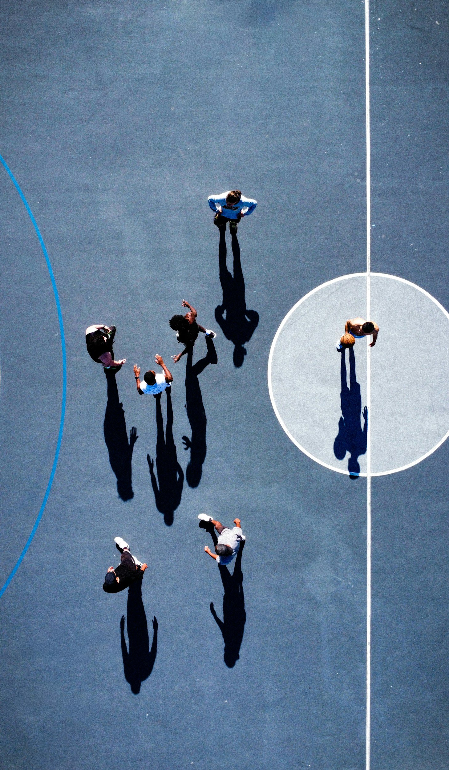 A group of people playing basketball on an outdoor court, viewed from above, with their shadows cast on the surface.