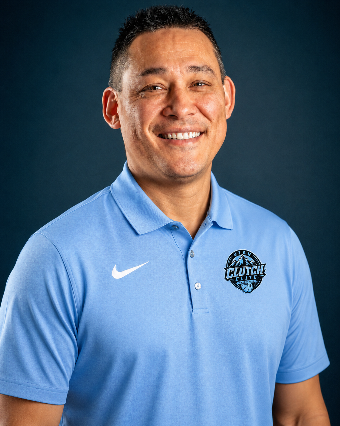 A smiling man wearing a light blue polo shirt with a Nike logo and a Utah Clutch Elite basketball logo, against a dark background.