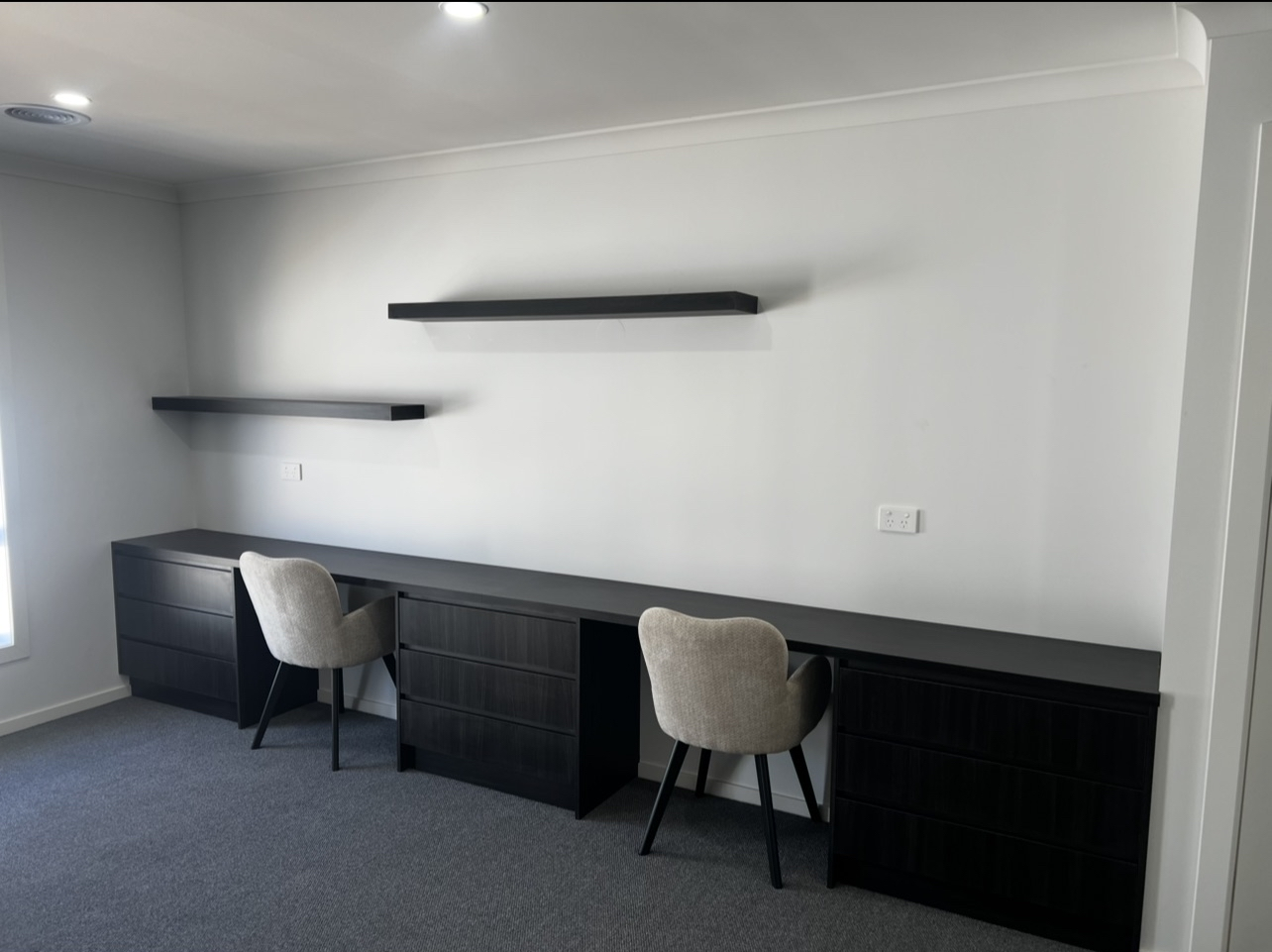 A modern home office with a long black desk, two gray chairs, and two floating black shelves on a white wall.
