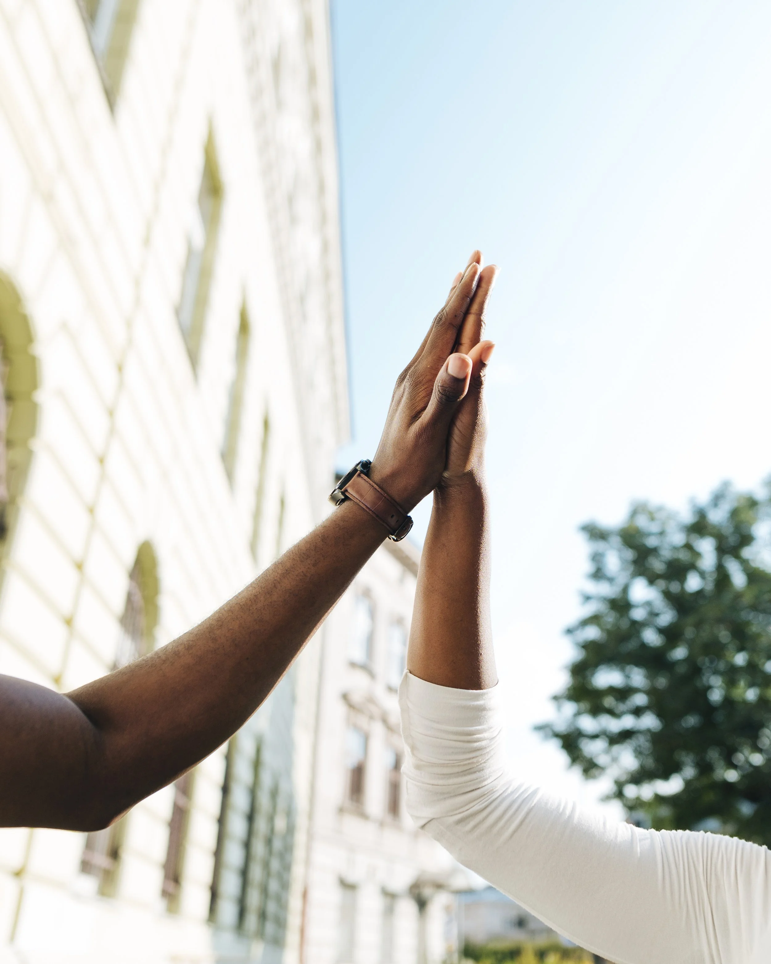 Two people reaching upwards to hold hands outdoors with a building and trees in the background.