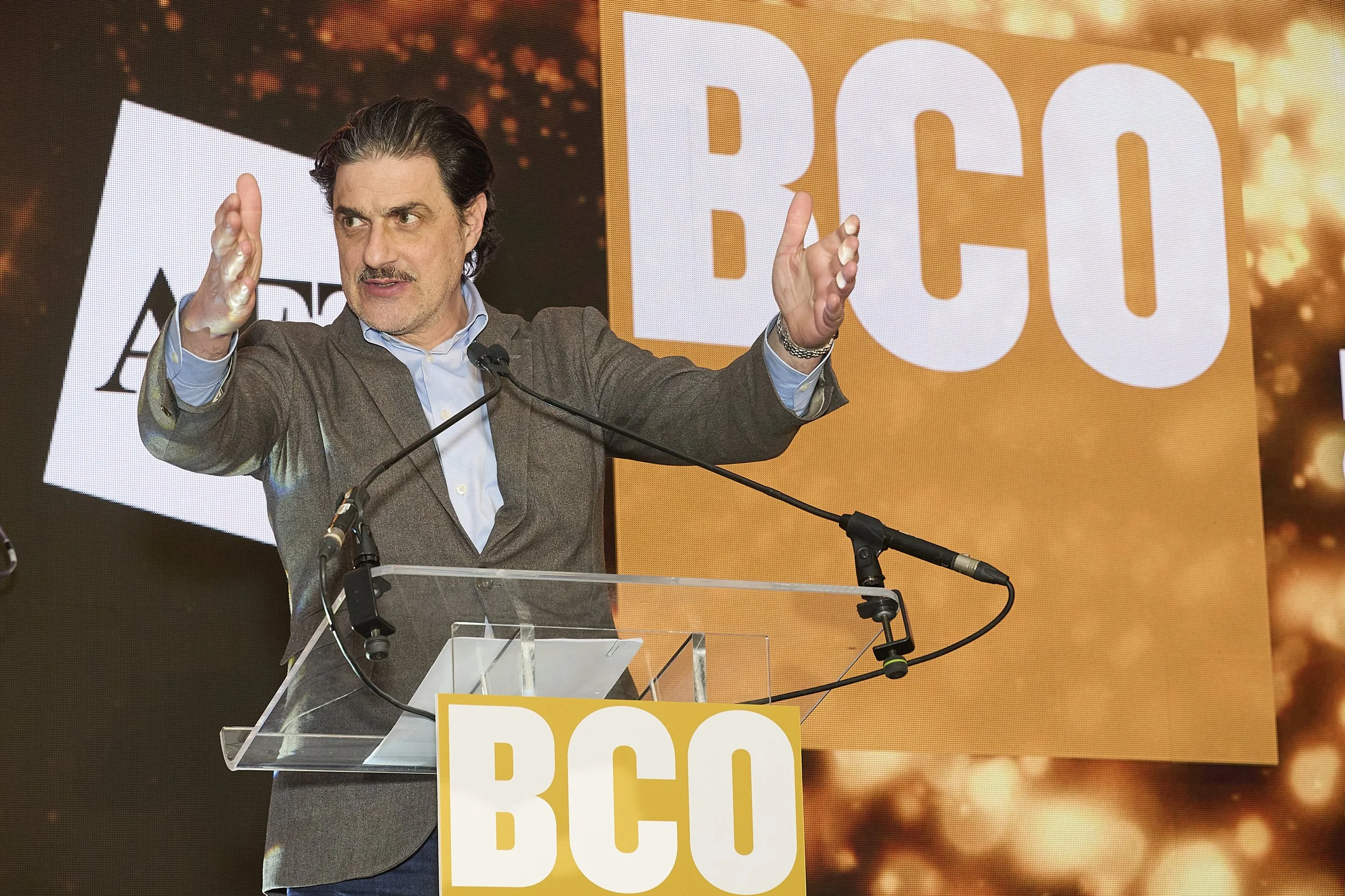 A man with dark hair and a mustache speaking at a podium during a BC Conservative event, with large yellow and white signs and a background with orange and yellow tones.