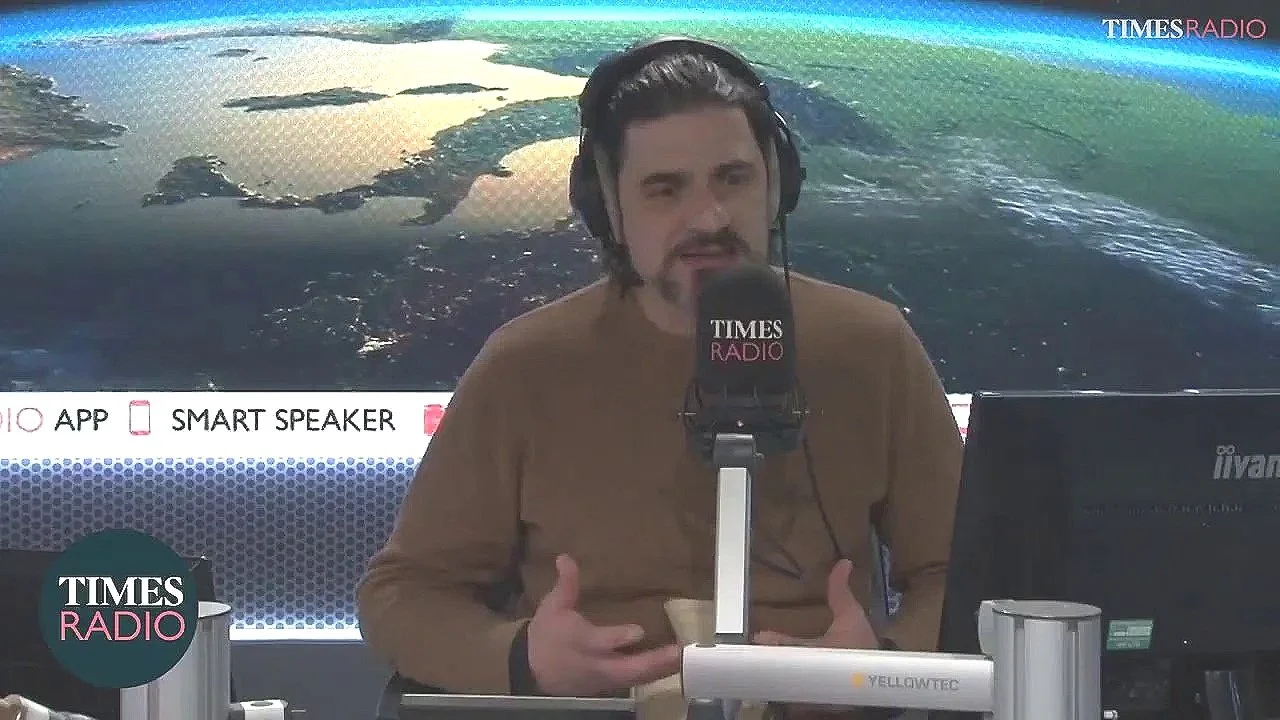 Alexis Conran into a TIMES RADIO microphone during a broadcast. He is wearing headphones and sitting in front of a computer, with a digital background of satellite imagery or a map behind him.