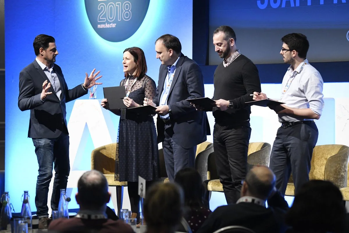 Alexis Conran on stage at a conference, engaged in conversation. Some audience members are holding notebooks or documents, with a large screen behind them displaying '2018 Manchester.' Audience members are seated in front, watching the event.