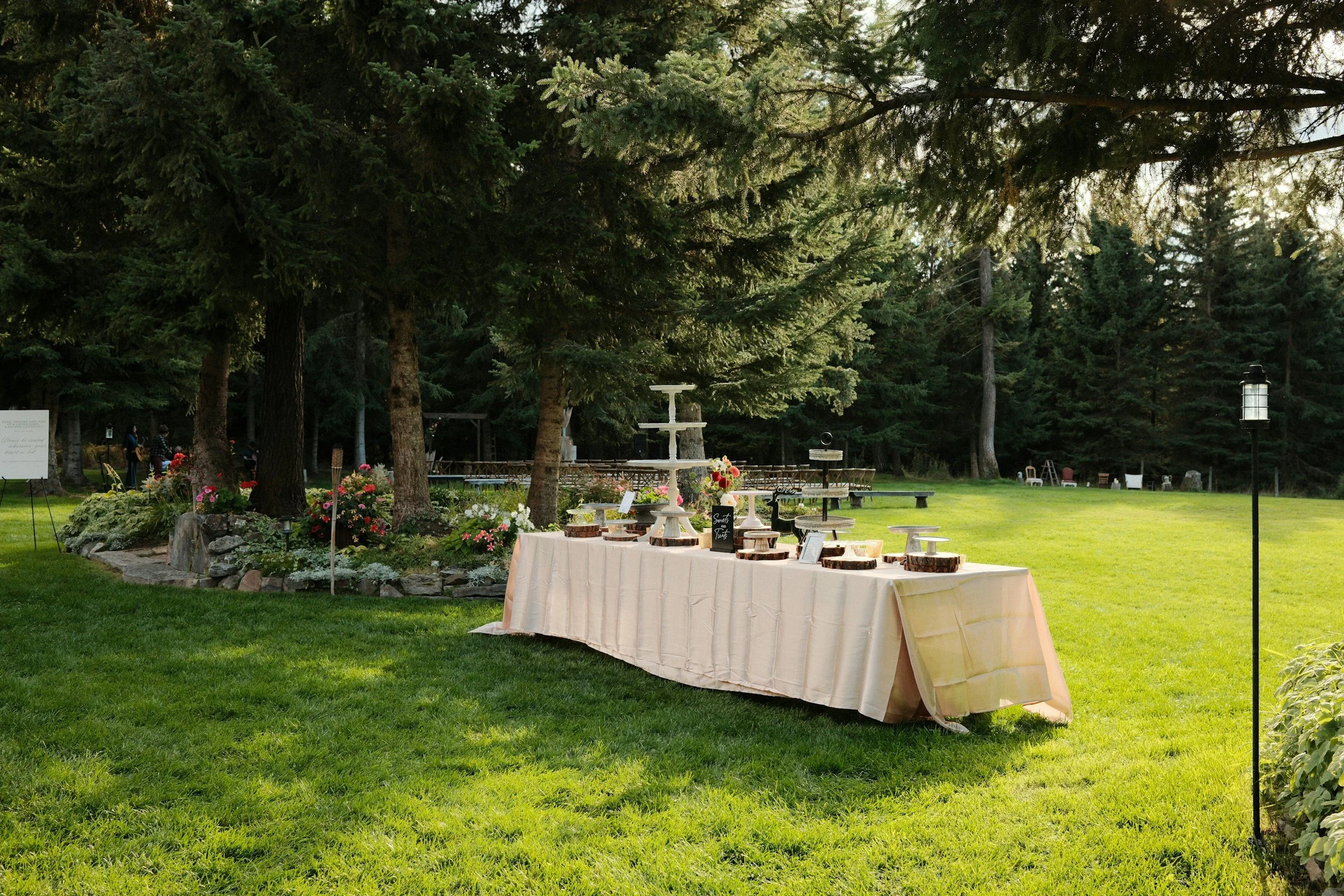 Outdoor garden setting with a table draped in a white cloth, decorated with flowers, fairy garden, and wood slices, surrounded by trees and lush greenery.