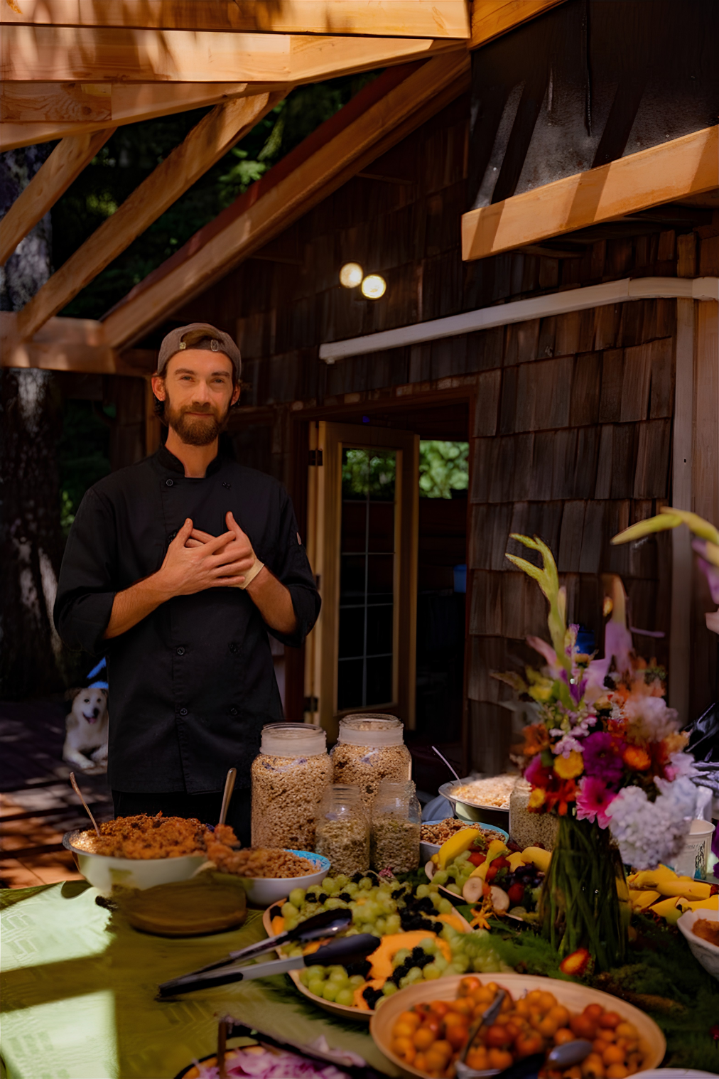A chef standing behind a table of assorted food and fruit, with a flower arrangement, outside a wooden building, with a dog sitting in the background.