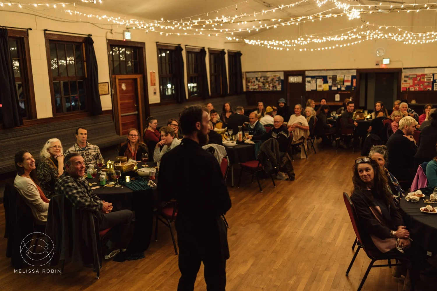 People seated at round tables in a decorated event hall listening to a speaker.