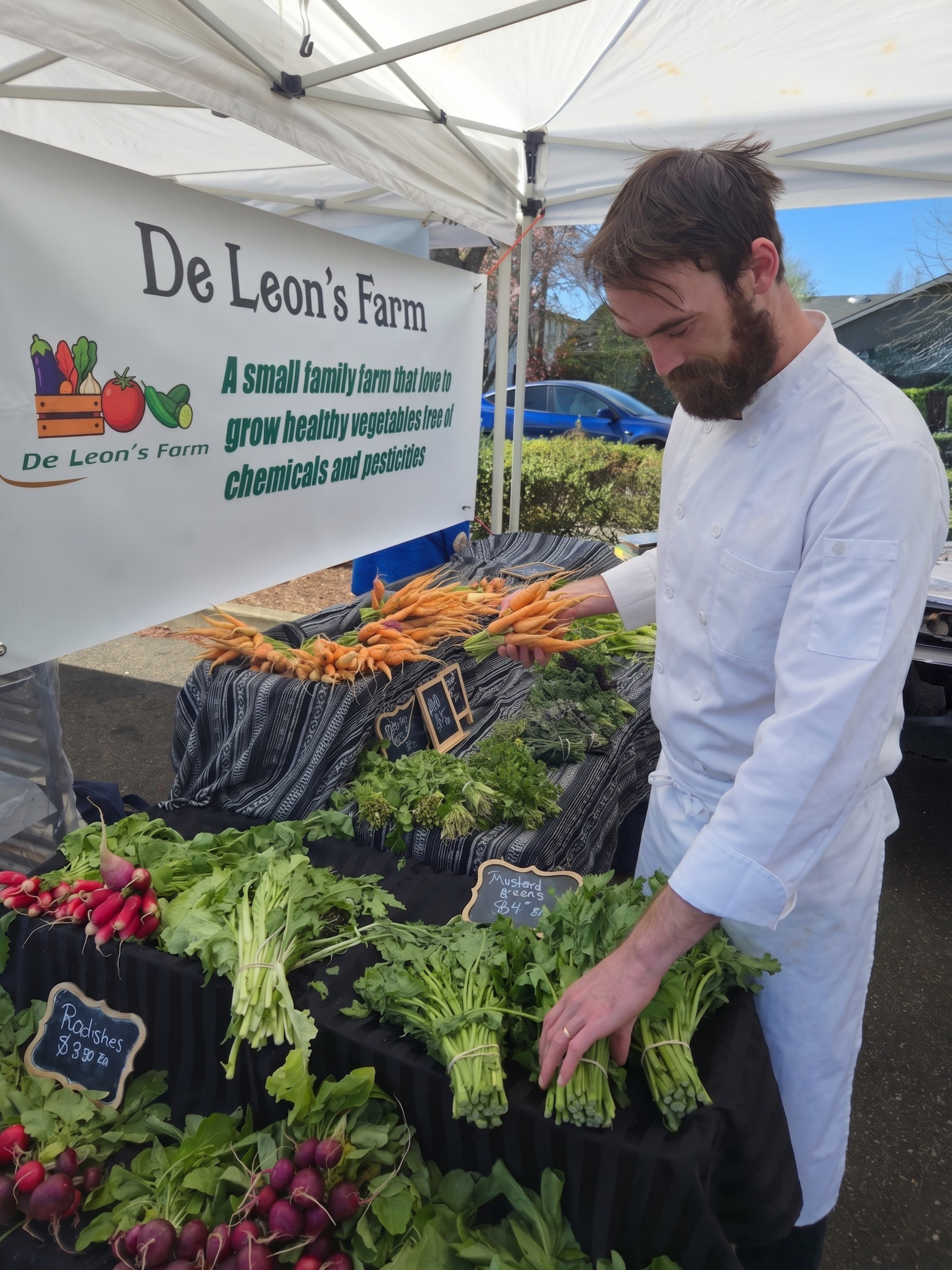 A man in a white chef uniform shopping for vegetables at a small farm stand titled 'De Leone's Farm'. The stand displays carrots, radishes, and leafy greens with small chalkboard signs.