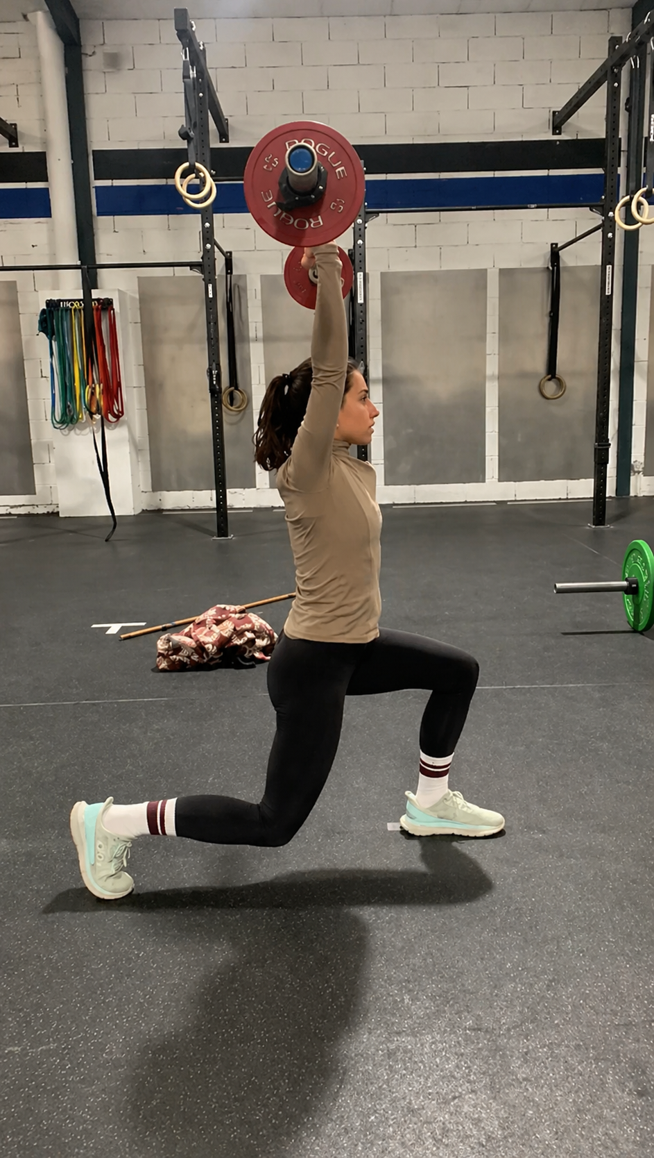 Une femme effectuant un exercice de musculation en salle de sport, tenant une barre avec des poids au-dessus de la tête, en position de fente."