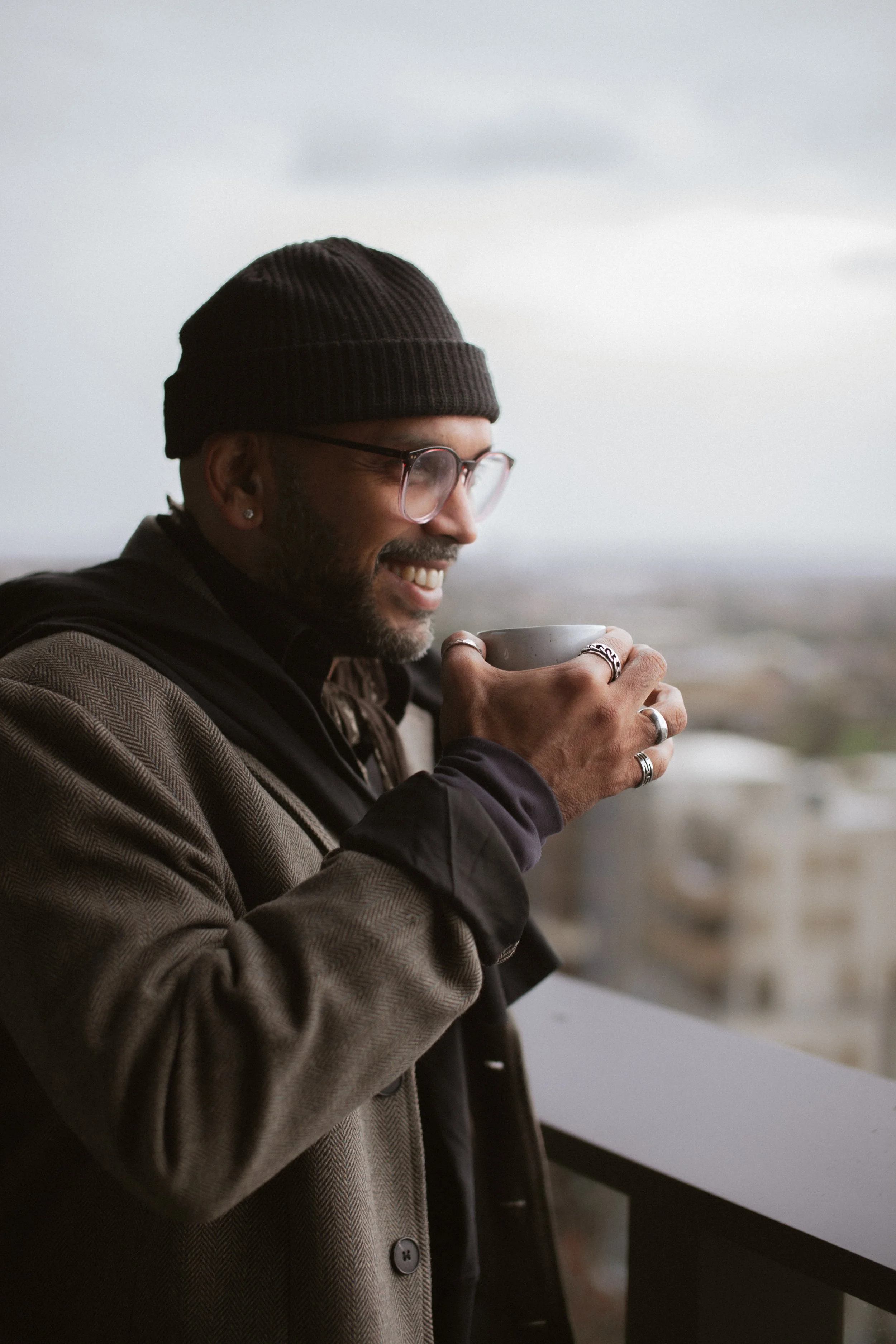 A man with glasses, earrings, and rings is smiling while holding a cup, wearing a beanie and a blazer, outdoors during daytime.