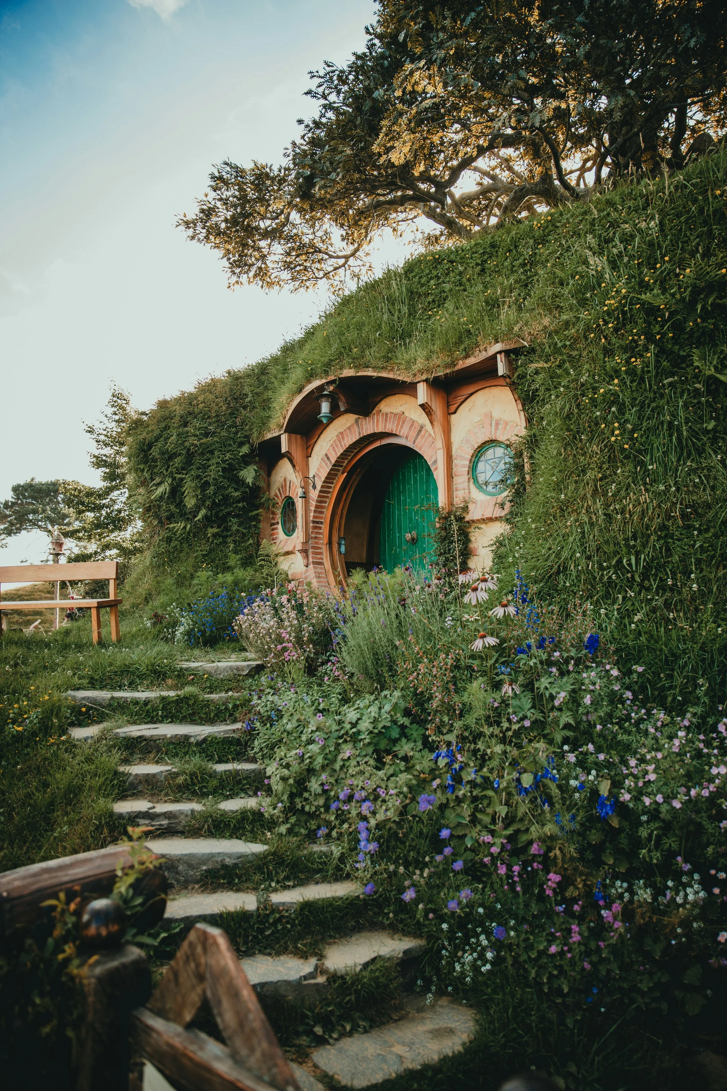 Entrance to a hobbit-style house built into a grassy hillside, with a green door and round windows, surrounded by colorful flowers and a stone pathway.