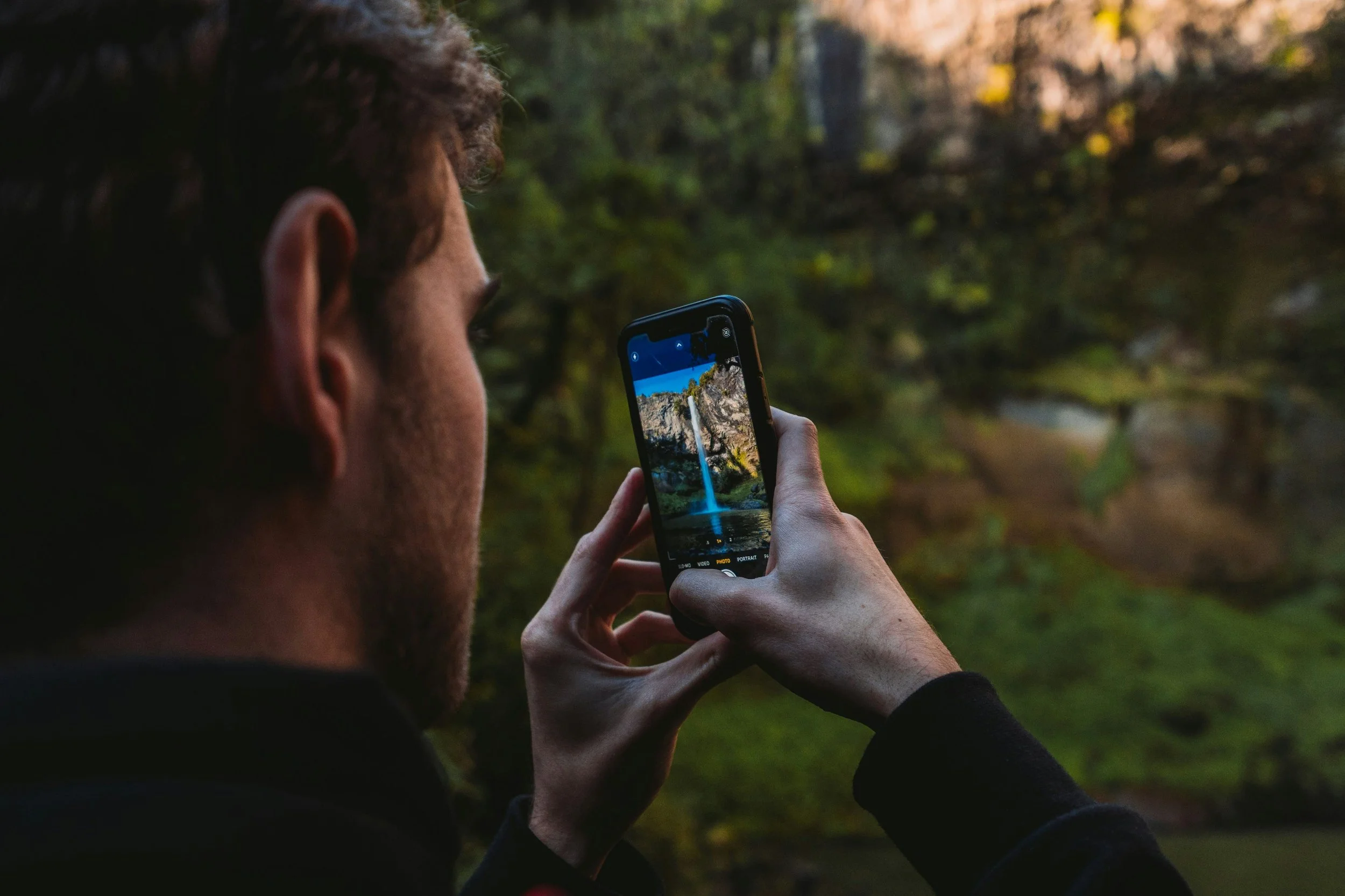 A person with curly hair taking a photo of a waterfall with a smartphone in a forest.