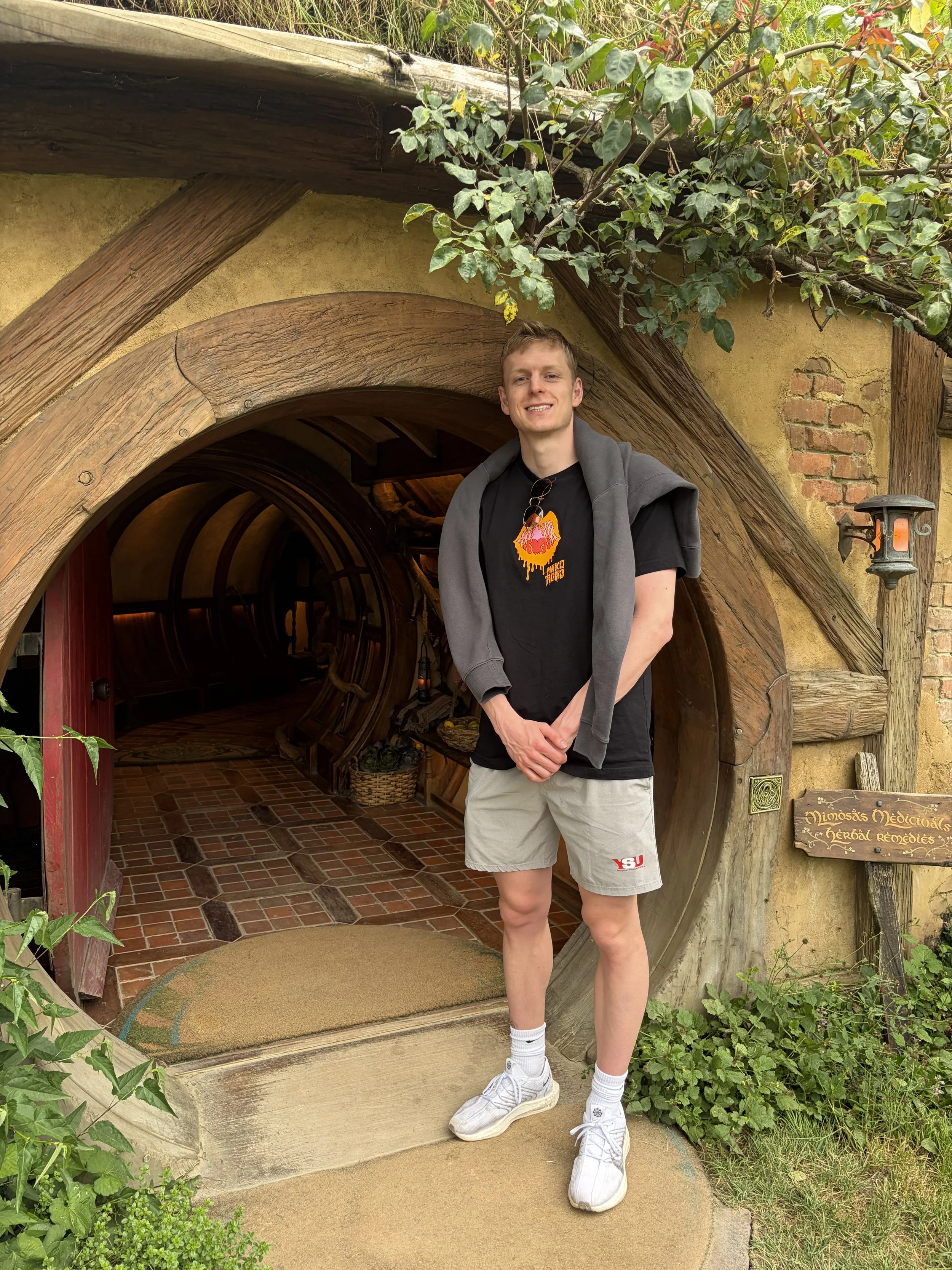 A young man standing outside a Hobbit-style house entrance with a rounded wooden door and brick path. He is smiling, wearing a black T-shirt, khaki shorts, white socks, and white sneakers, with a gray sweatshirt draped over his shoulders. The house has a rustic, whimsical appearance with wood and brick details, surrounded by greenery.
