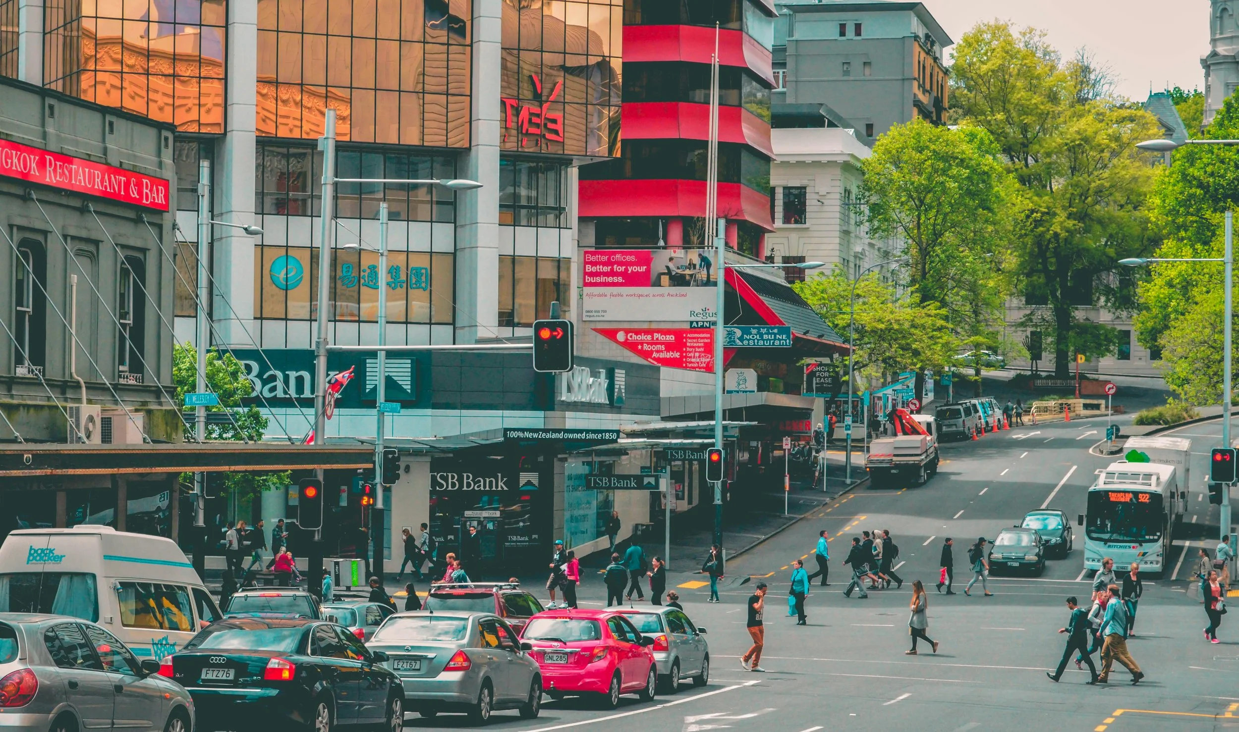 City street scene with cars, buses, and pedestrians. Tall buildings with advertisements and signs, including TSB Bank and a restaurant. Green trees lining the street, traffic lights, and people crossing at the intersection during daytime.