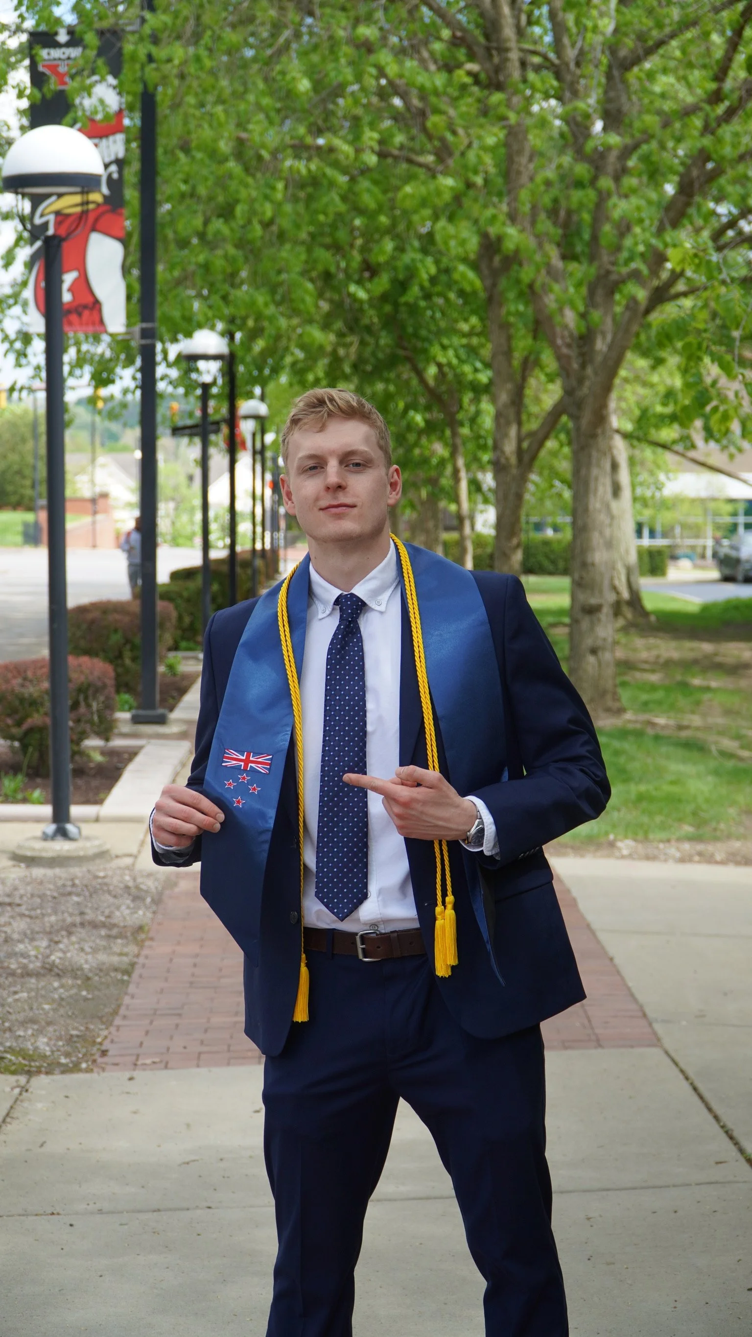 Young man in a graduation cap and gown pointing at his diploma, standing outdoors on a sidewalk with trees and streetlights in the background.
