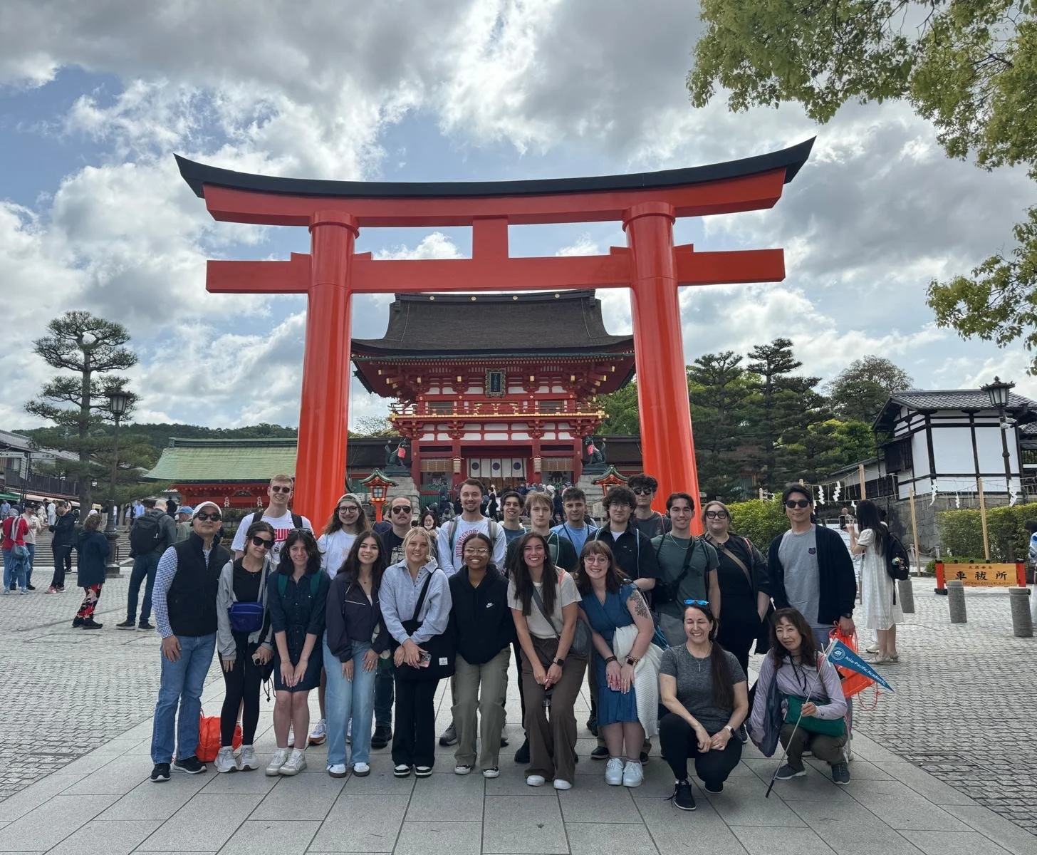 Group of tourists standing in front of a large red torii gate at a traditional Japanese shrine on a partly cloudy day, with multiple buildings and trees in the background.