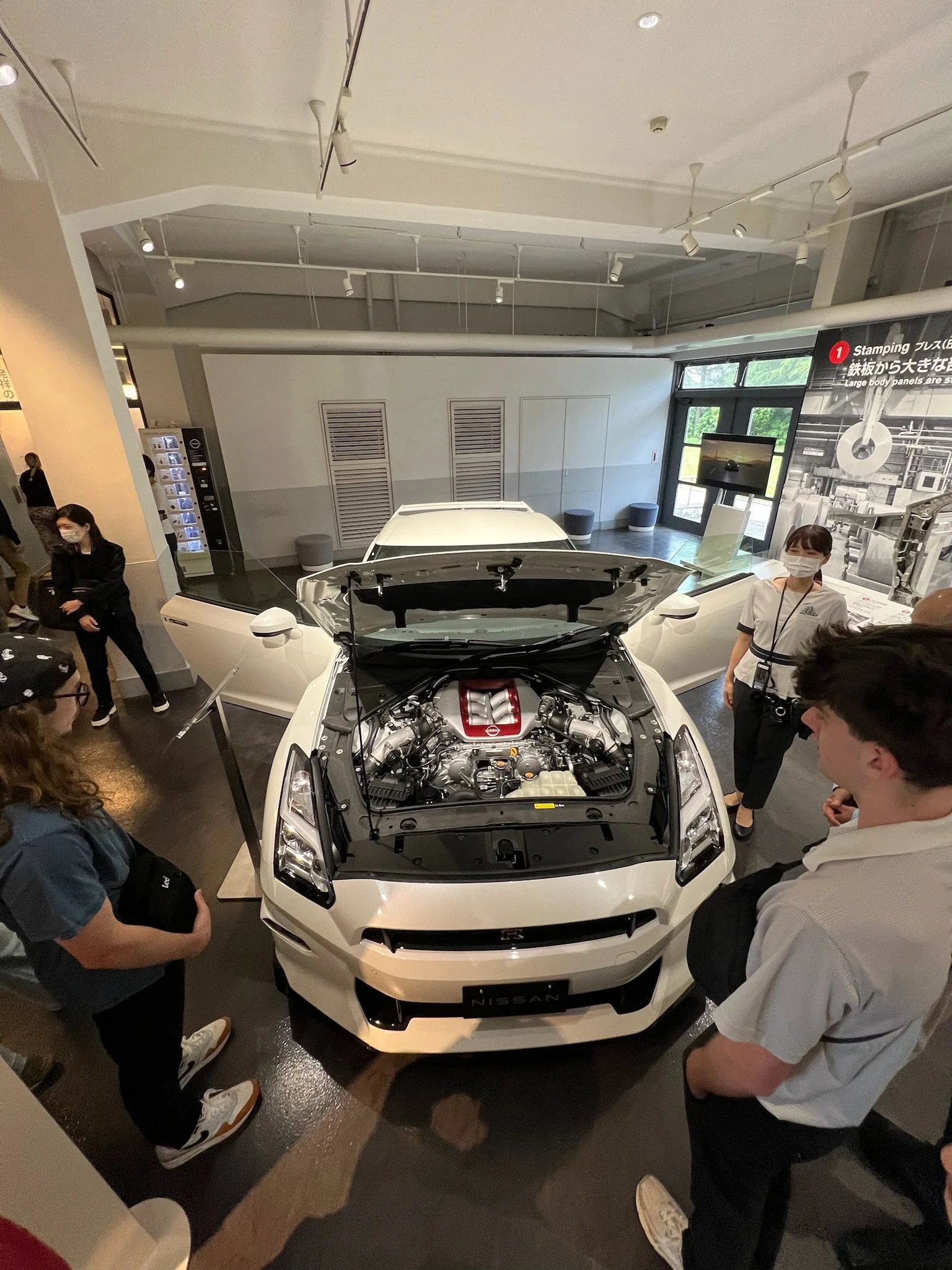 A group of people gathered around a white Nissan sports car with its hood open, displaying the engine. The setting appears to be an indoor showroom or exhibition space, with some people wearing masks and others taking notes or looking at the car.