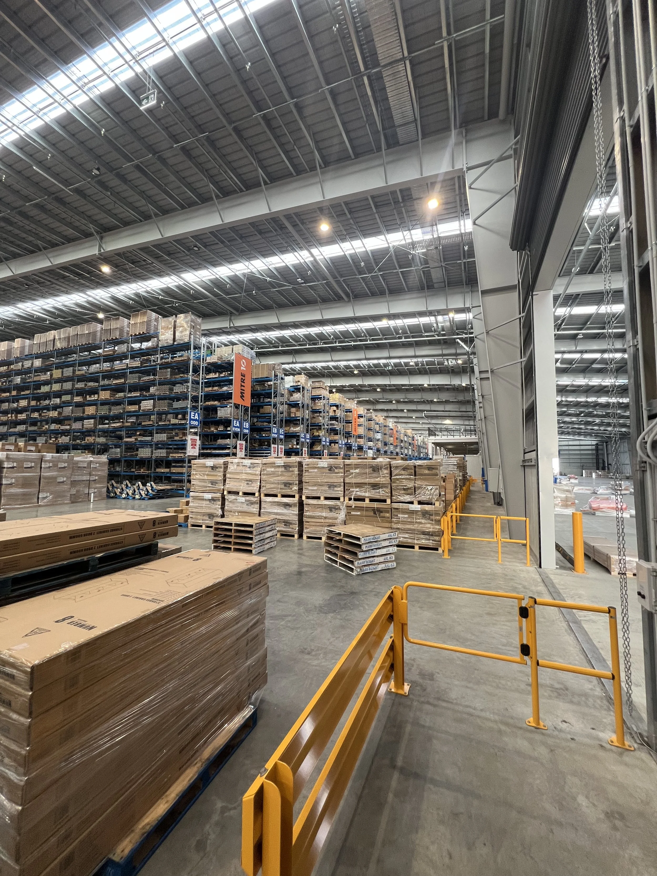 Interior of a warehouse with shelves filled with boxes and pallets, and safety barriers in the foreground.