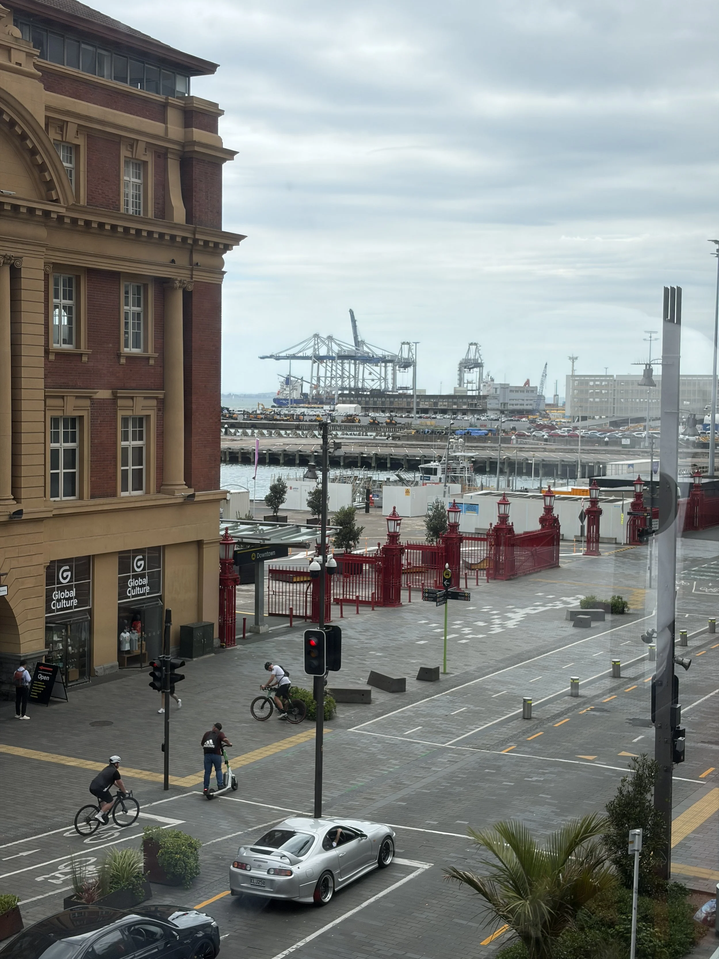 View of a waterfront area with a historic building on the left, people biking and walking on the street, a parking lot with cars, and a harbor with cranes and ships in the background under cloudy sky.