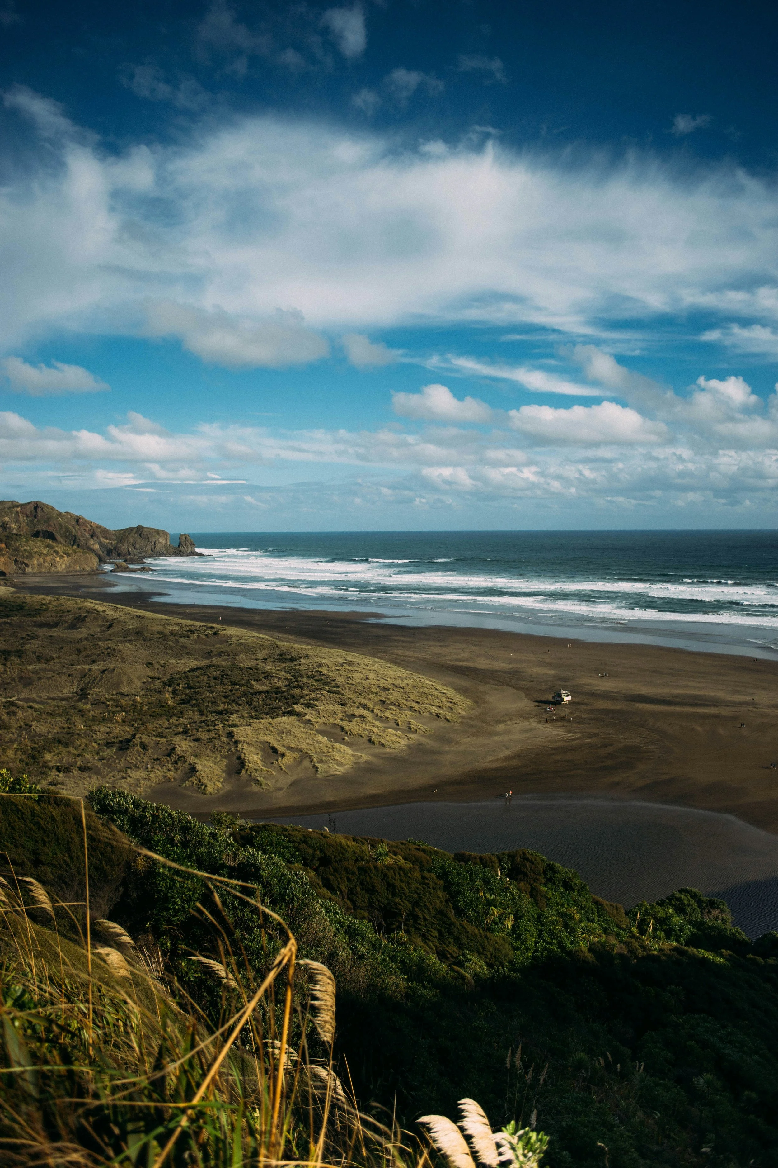 A coastal landscape featuring a beach, waves, cliffs, and a partly cloudy sky with patches of blue.