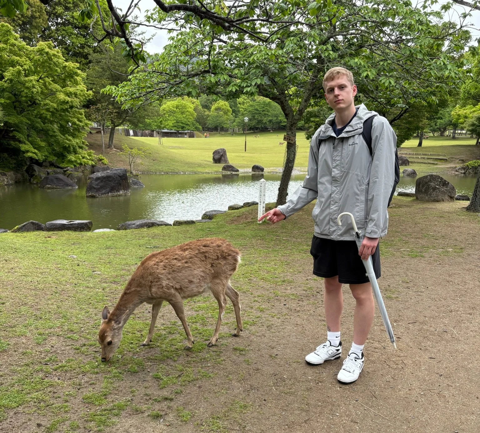 A young man with a backpack and crutches standing near a small animal, possibly a baby goat, by a pond in a park with lush greenery and trees.