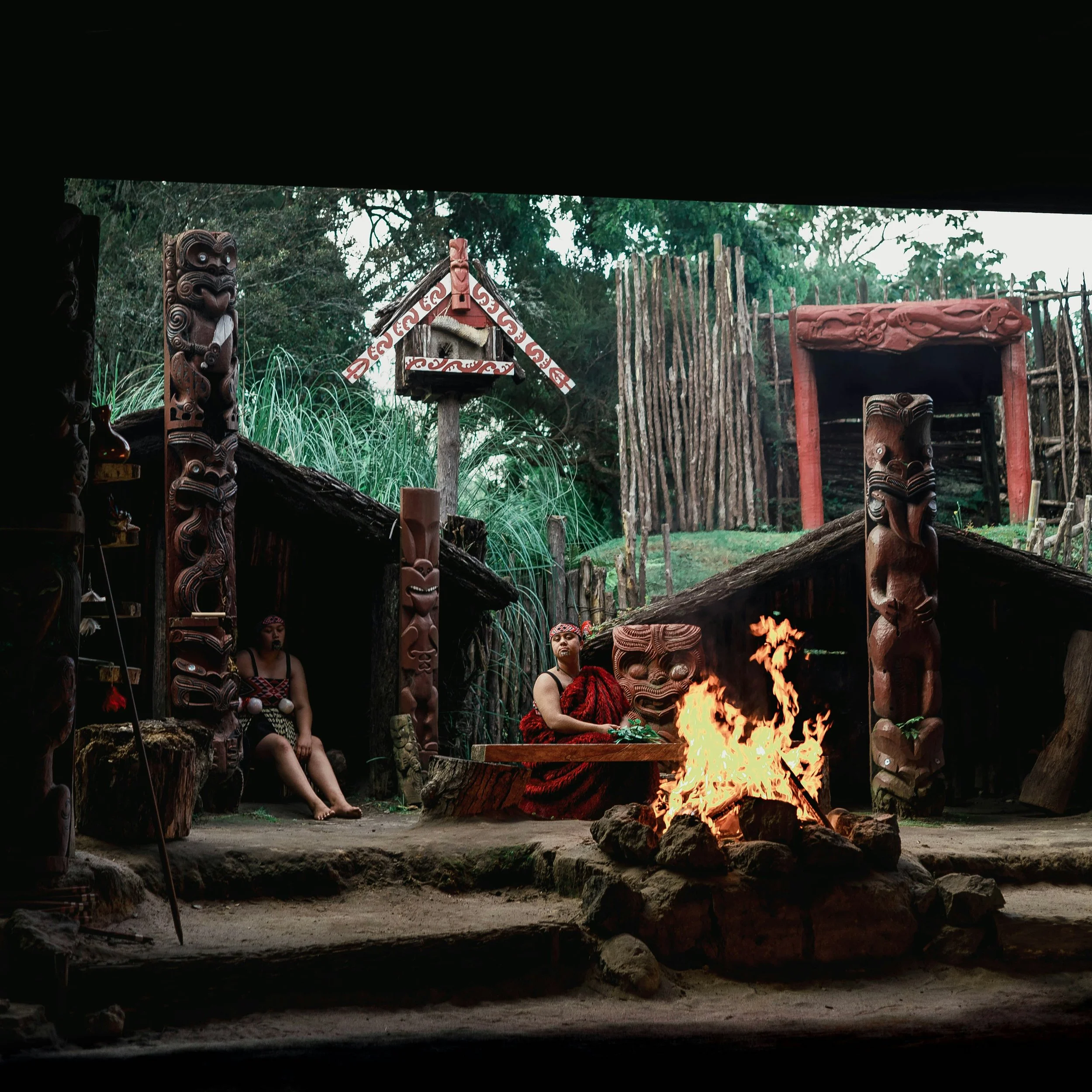 A traditional Maori hangi and cultural scene with carved wooden figures, a fire pit, and people in tribal attire, set outdoors in a lush green environment.