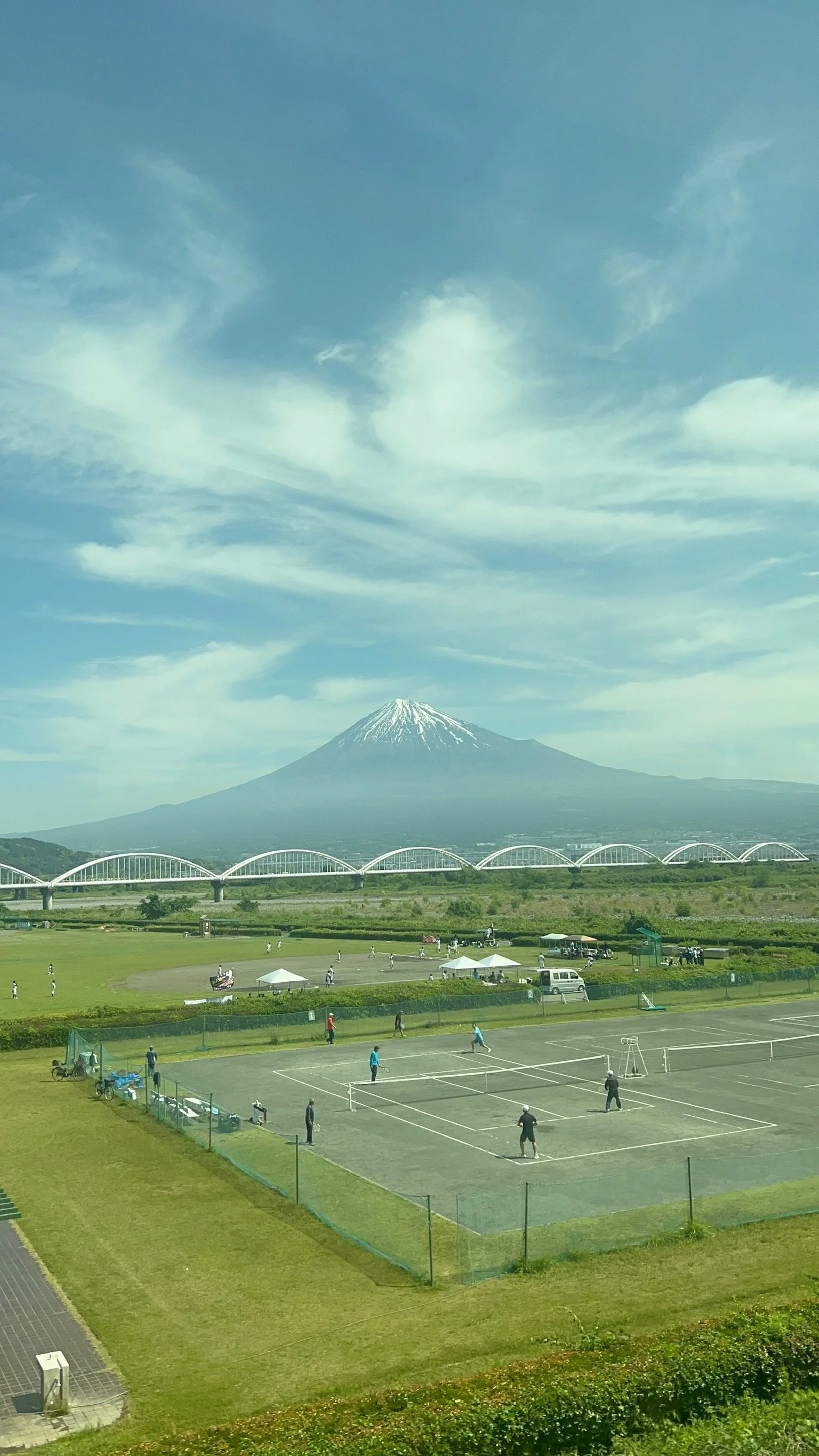 A scenic view of Mount Fuji with snow-capped peak, a park in the foreground with people playing tennis, walking, and relaxing, and a bridge crossing a valley beneath the mountain under a partly cloudy sky.