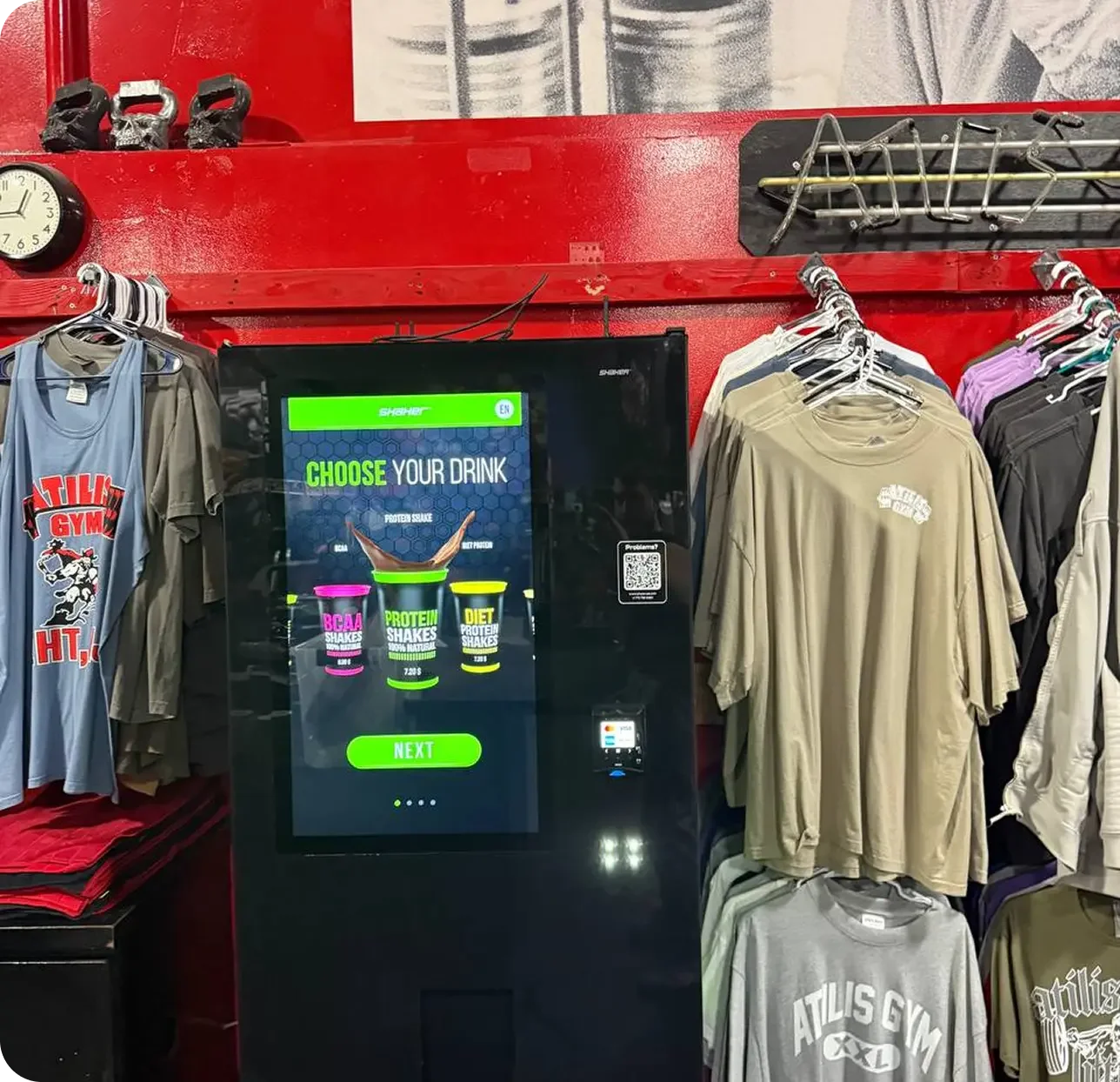 Un distributeur automatique pour choisir une boisson protéinée, entouré de vêtements de sport suspendus dans une boutique.