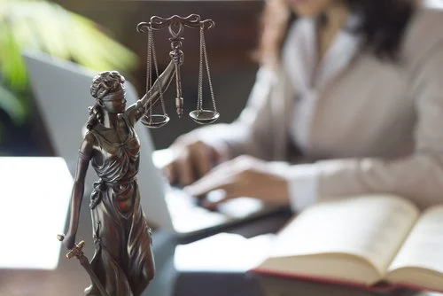 Close-up of a bronze Lady Justice statue with scales, in front of a person working on a laptop and an open book.