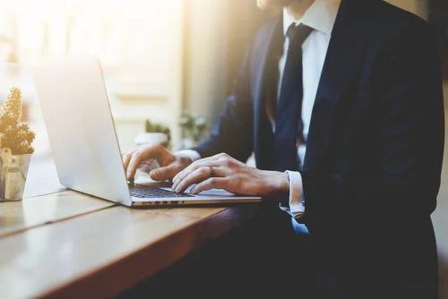 Person in business attire working on a laptop at a wooden desk in a bright office.