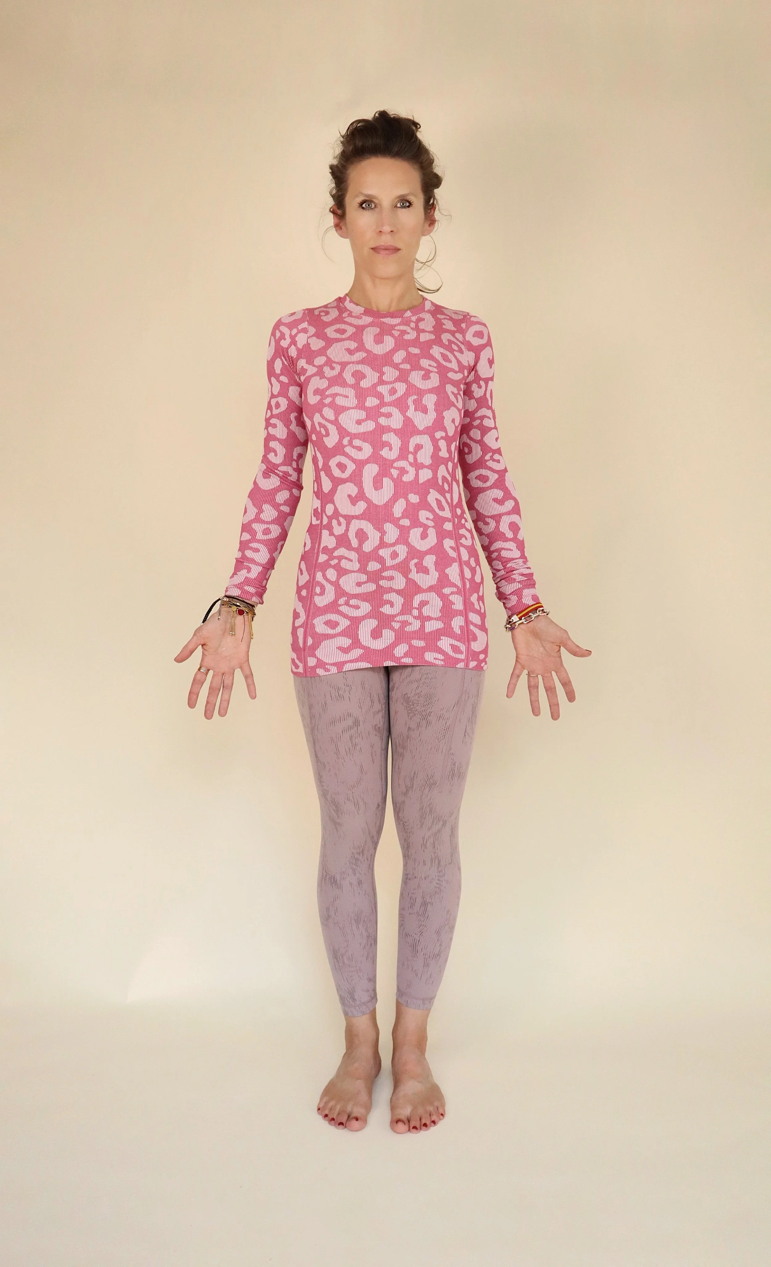 Woman standing barefoot against a plain beige background, wearing a pink patterned long-sleeve top and light pink patterned pants, with arms slightly extended and hands open.
