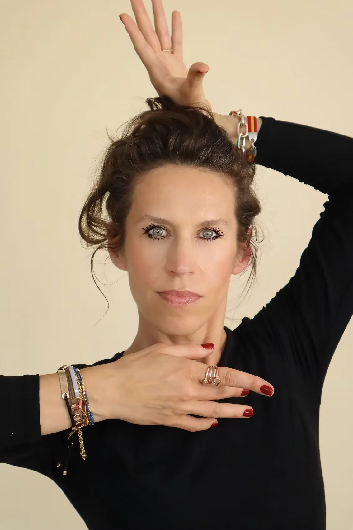 A woman with blue eyes and brown hair in a black top, posing with her hands in a ballet-like position, one above her head and one below her face, against a beige background.