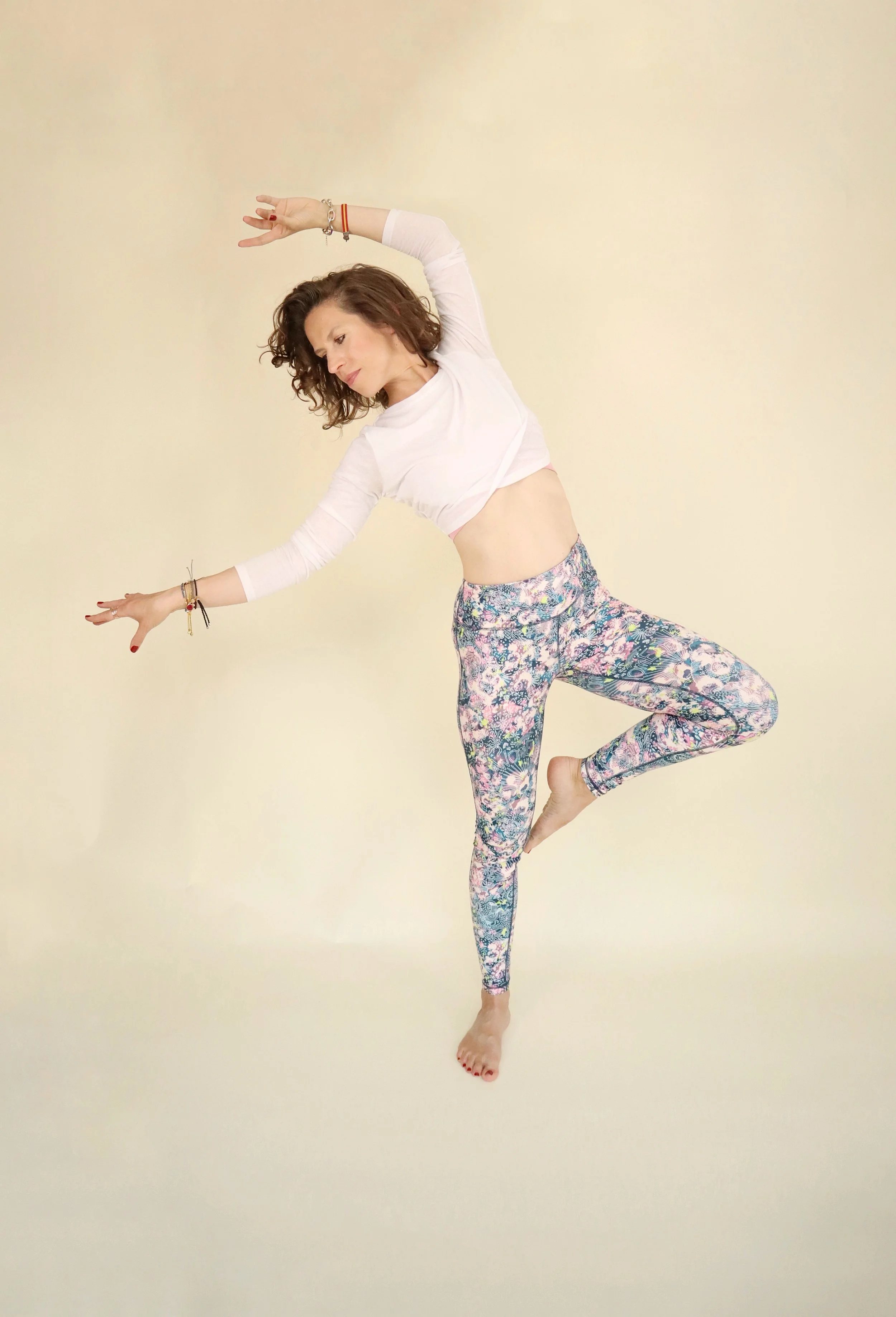 A woman practicing yoga in a balancing pose on a light-colored background, wearing a white top and colorful patterned leggings.