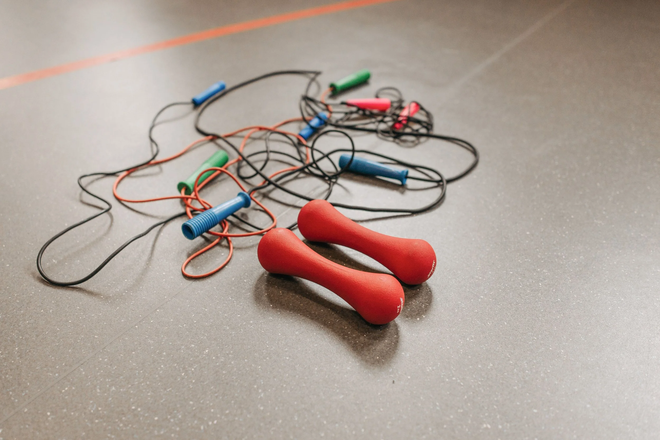 A pair of red massage rollers and tangled resistance wires and earphone wires on a gray floor.