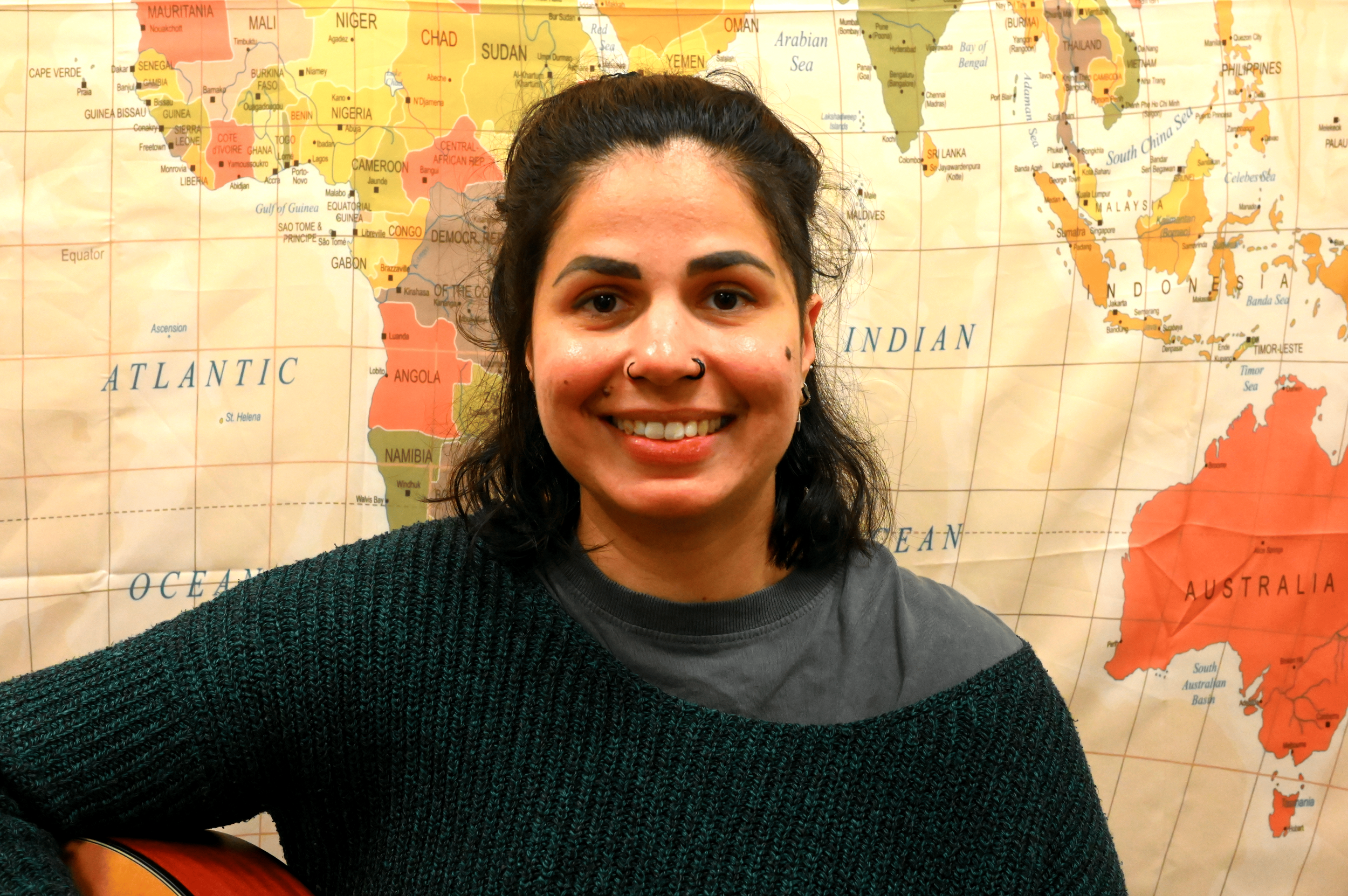A smiling woman with dark curly hair, a nose ring, and dark eye makeup, wearing a gray shirt and a dark green sweater, standing in front of a colorful world map background.