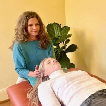 Cornelia Bühlmann bei kinesiologische Behandlung am Kopf bei einer Frau die auf einer Liege liegt, in einem Praxisraum mit gelben Wänden und einer grossen grünen Pflanze im Hintergrund.