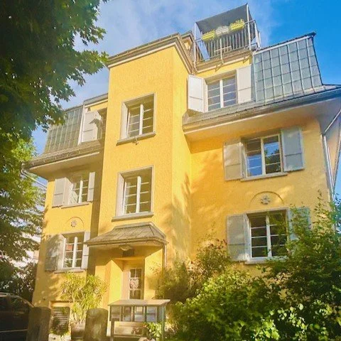 Villa Widnau ein gelbes mehrstöckiges Gebäude grünen Fensterläden, Balkonen und Dachterrasse, umgeben von Bäumen, bei Tageslicht.