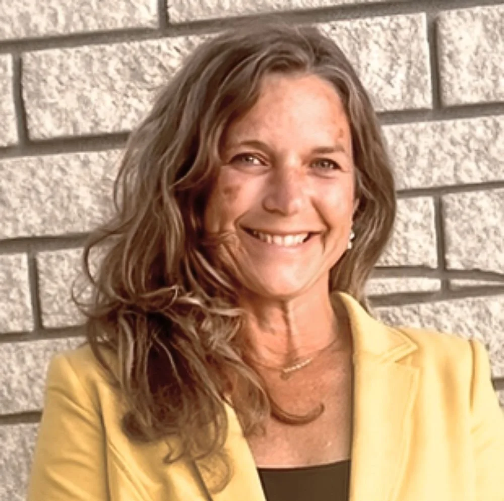 Cornelia Bühlmann mit braunen, lockigen Haaren trägt eine gelbe Jacke und steht lächelnd vor einer Backsteinmauer.
