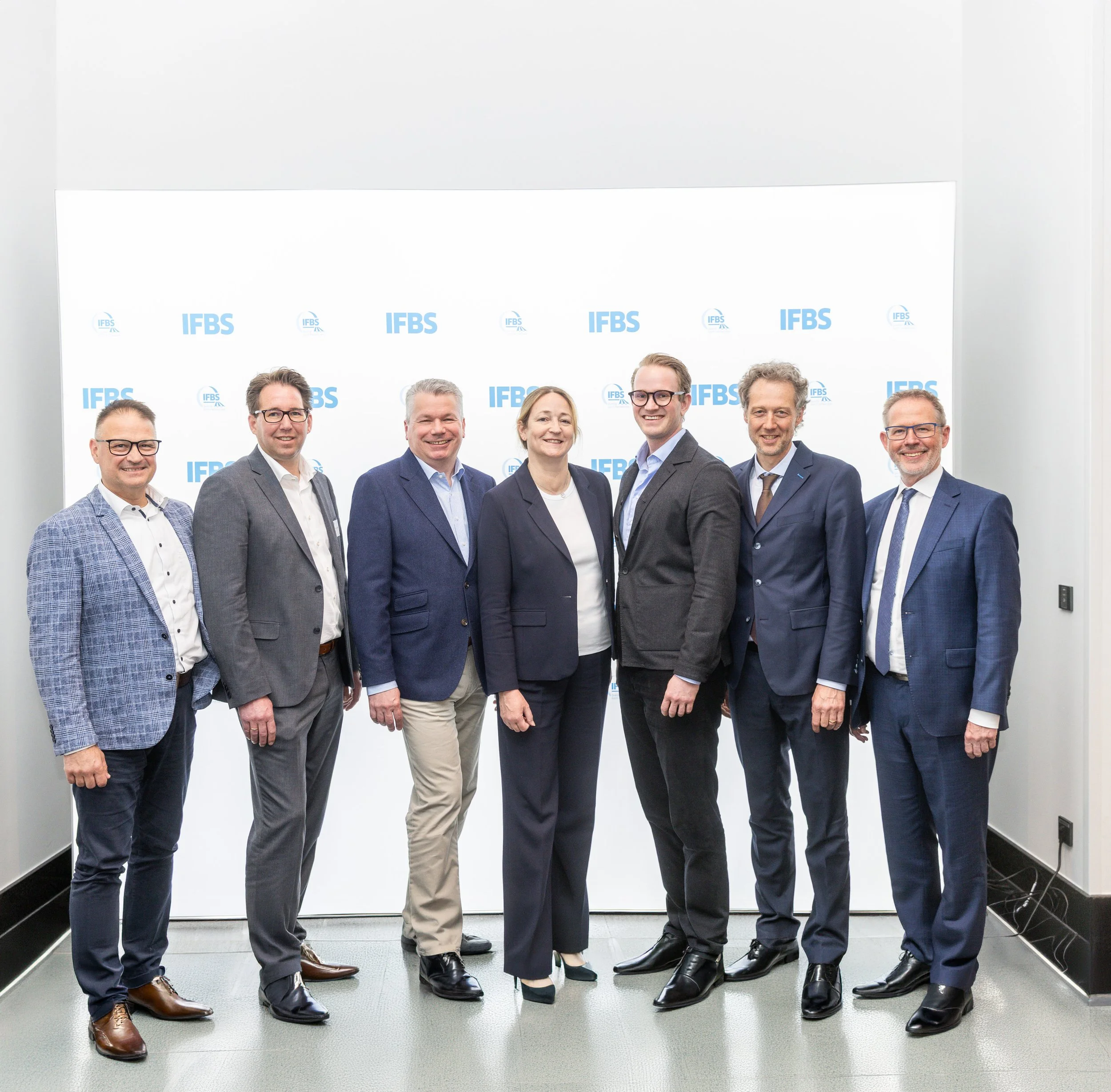Eine Gruppe von acht professionell gekleideten Menschen, sechs Männer und zwei Frauen, posieren vor einem weißen Hintergrund mit dem Logo 'IFBS'.
