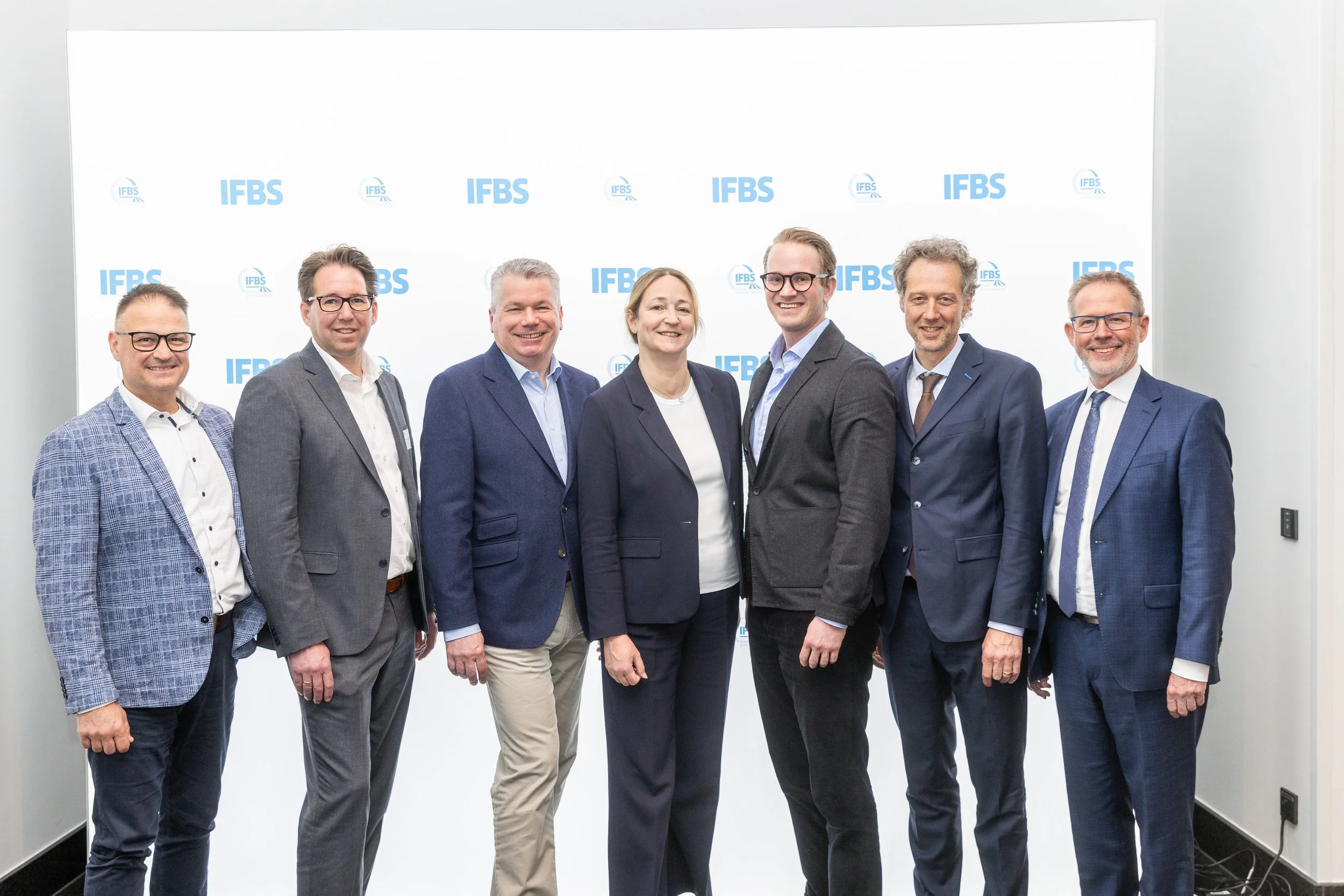 Gruppenfoto mit sieben Personen in formeller Kleidung vor einer weißen Wand mit den blauen Logos des IFBS.